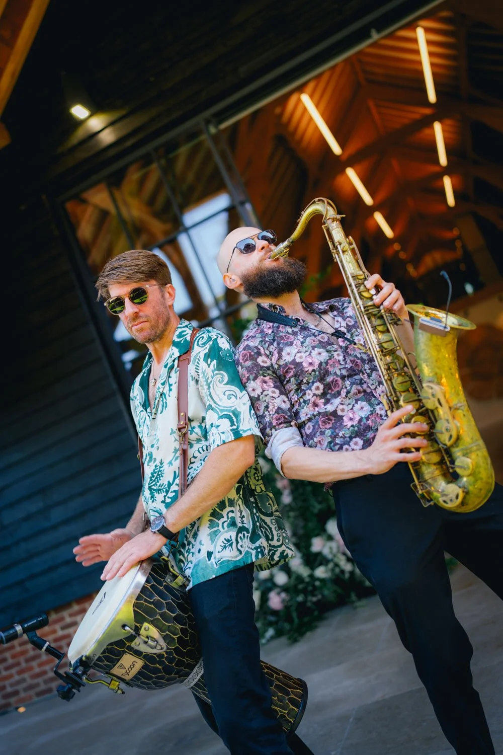 Two musicians wearing sunglasses perform with a drum and a saxophone in an indoor venue with wooden architectural details.
