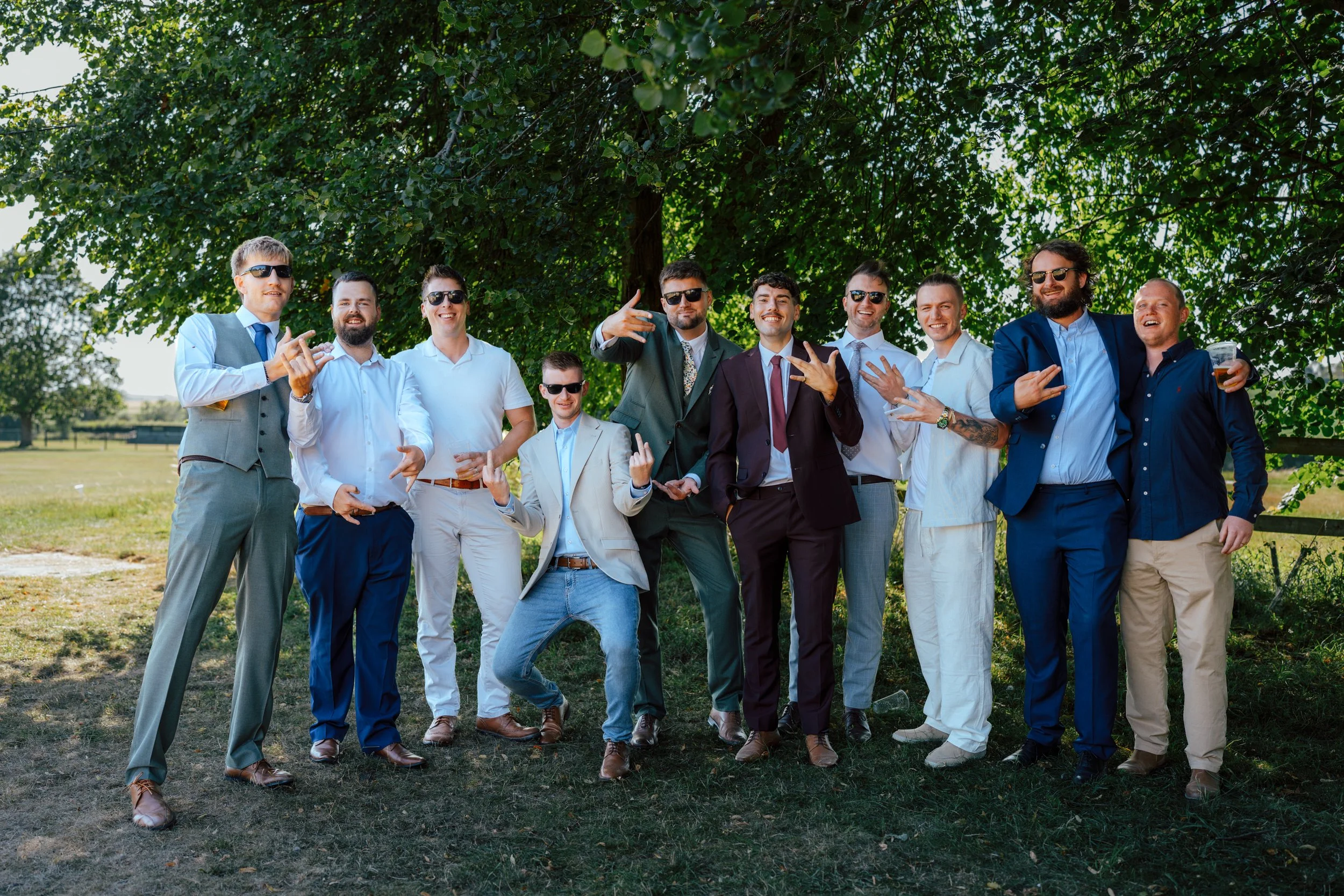 Group of men and women standing outdoors under a large tree, dressed in formal and semi-formal attire, some wearing sunglasses, smiling and posing for a photo.