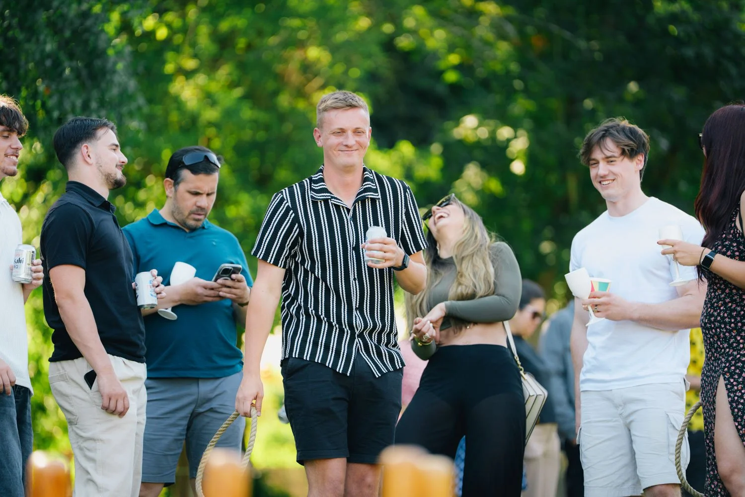 Group of people outdoors at a social gathering, smiling and holding drinks, with lush green trees in the background.