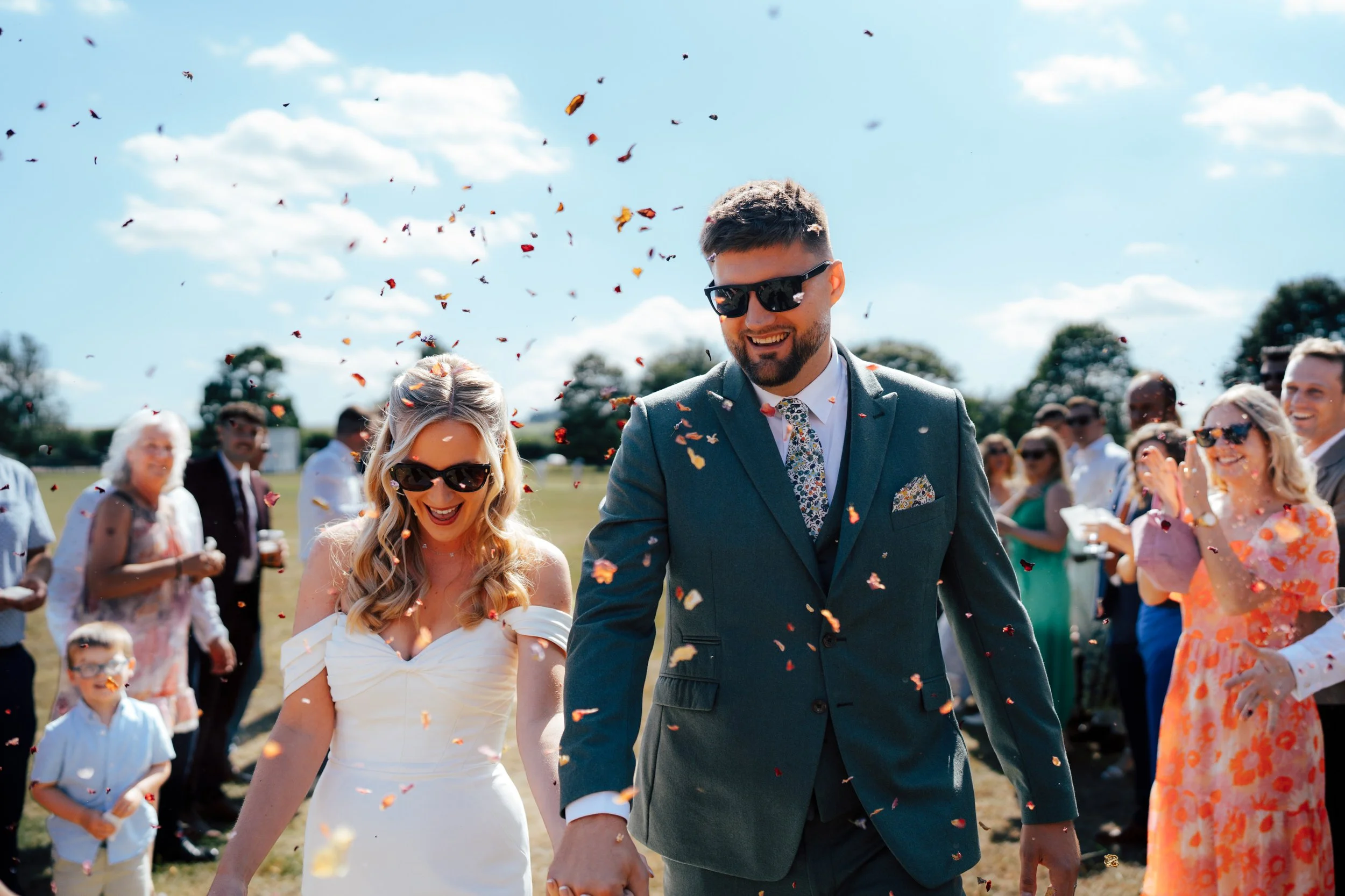 A couple dressed in formal wedding attire holding hands and smiling amidst a shower of colorful confetti, with guests in the background celebrating outdoors on a sunny day.