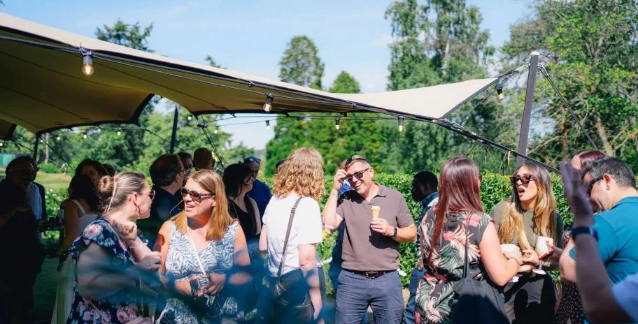 Guests mingling, eating ice cream, and laughing under a stretch canopy tent at a sunny outdoor summer party, candid event photography by Alex Gell.