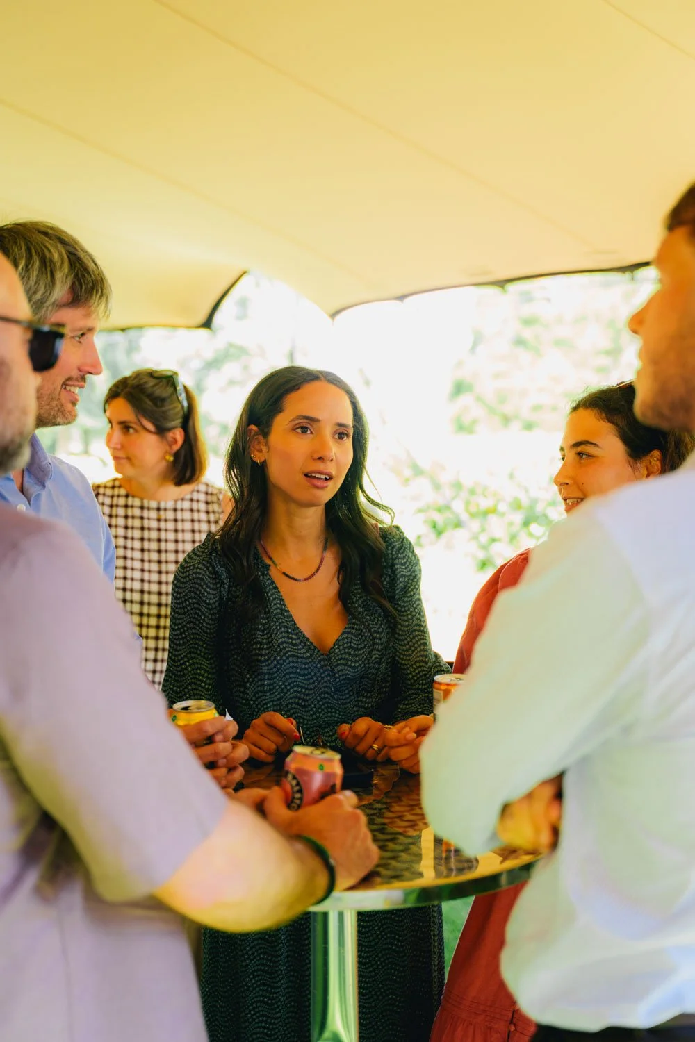 Group of people socializing outdoors, gathered around a table, enjoying drinks, with a woman in center speaking and others listening.