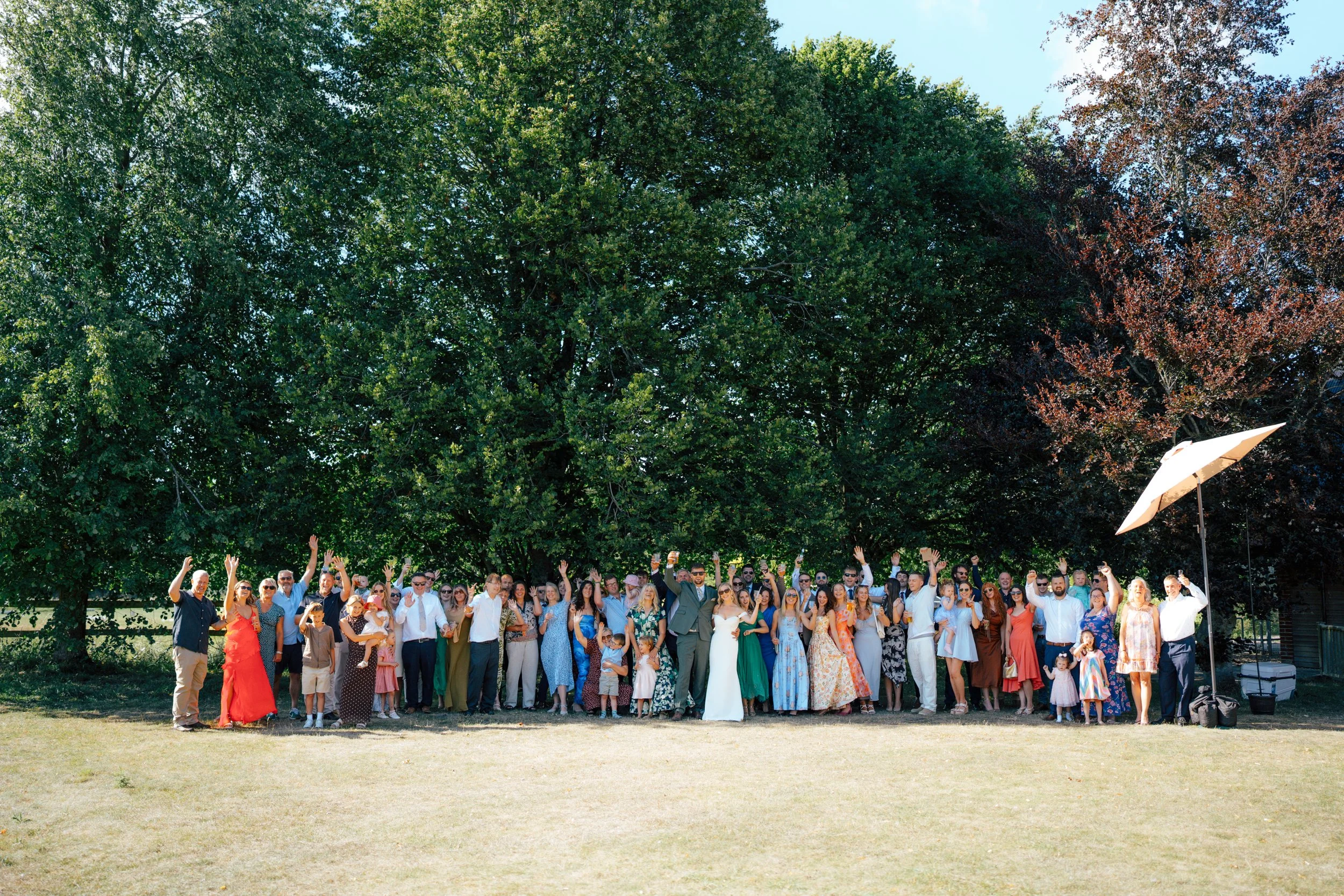 Large group of people gathered outdoors under tall green and reddish trees, celebrating or attending an event, with some raising hands and smiling.