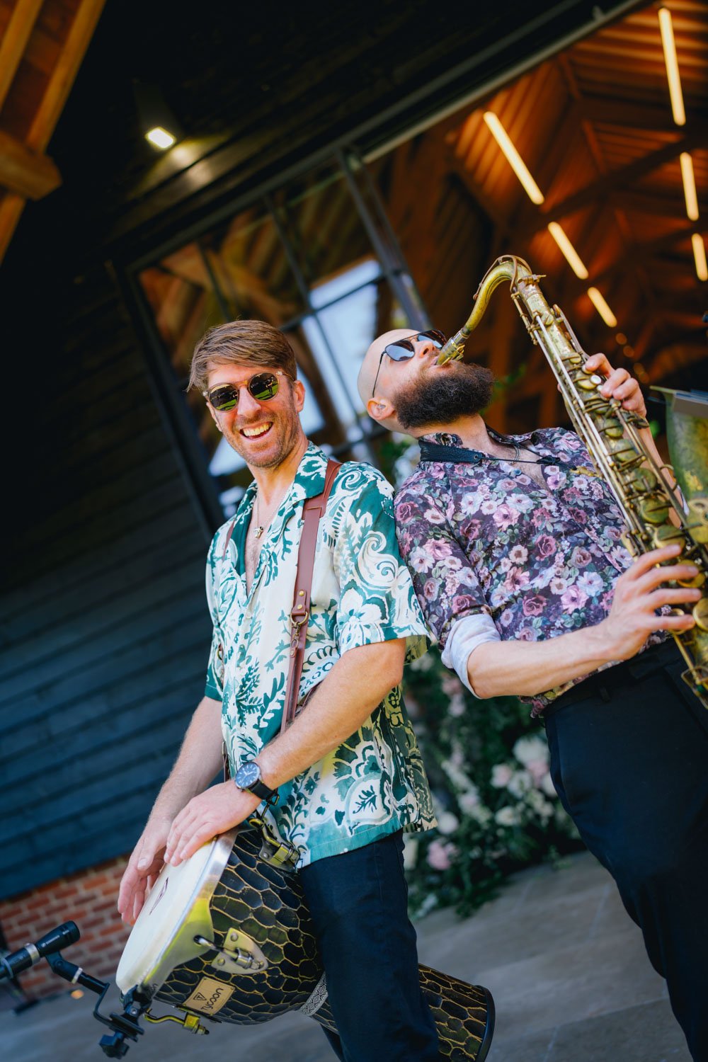 Two men playing musical instruments, one with a tambourine and the other with a saxophone, wearing sunglasses and colorful patterned shirts, smiling and enjoying themselves outdoors under a wooden shelter.
