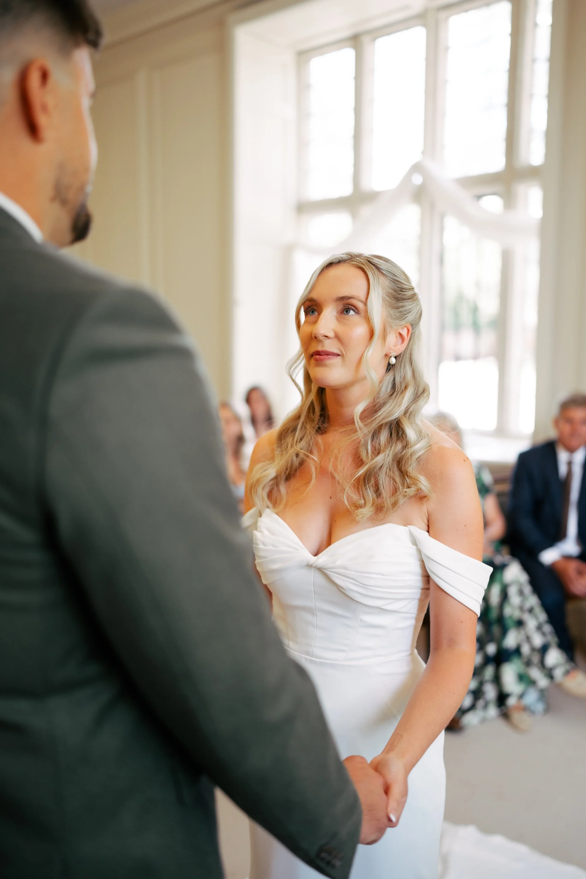 A bride and groom holding hands during their wedding ceremony in a bright room with large windows.