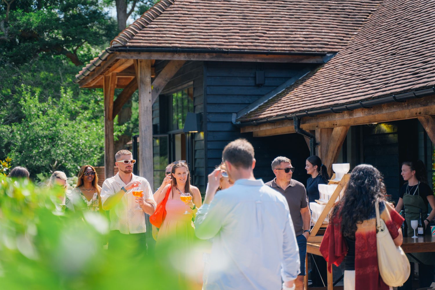 People socializing outdoors at a gathering near a dark wooden house with a tiled roof.