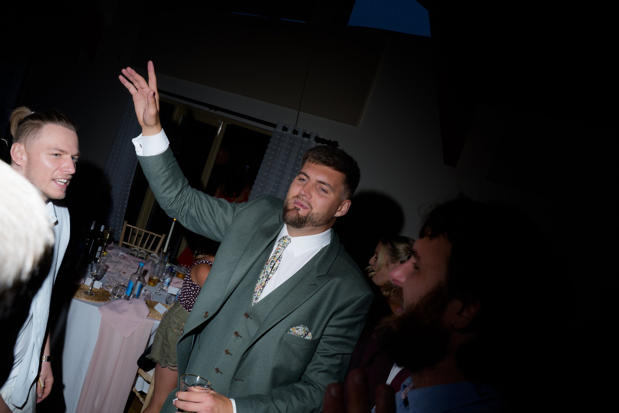 Two men at a social gathering, one raising his hand and the other holding a glass, with a table of drinks and people in the background.