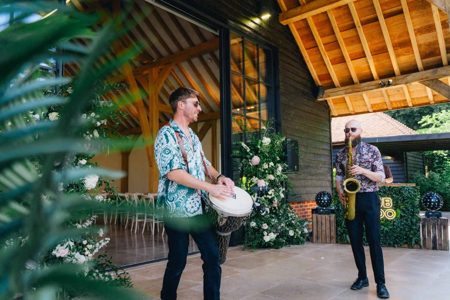 Two musicians playing instruments outdoors at a social event, surrounded by greenery and flowers, with a wooden building in the background.
