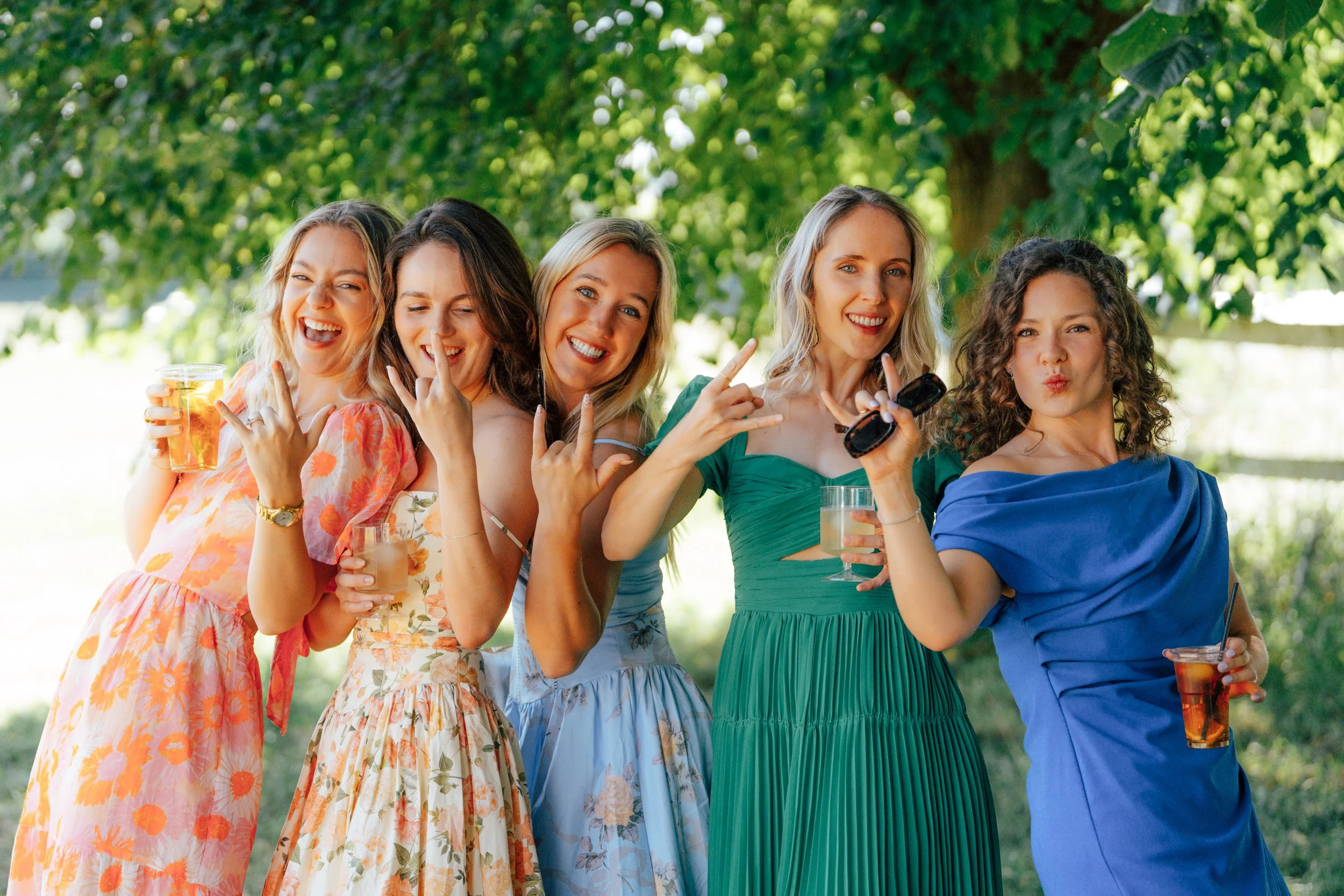 Group of six women standing outdoors under a green tree, smiling and holding drinks, dressed in colorful summer dresses.