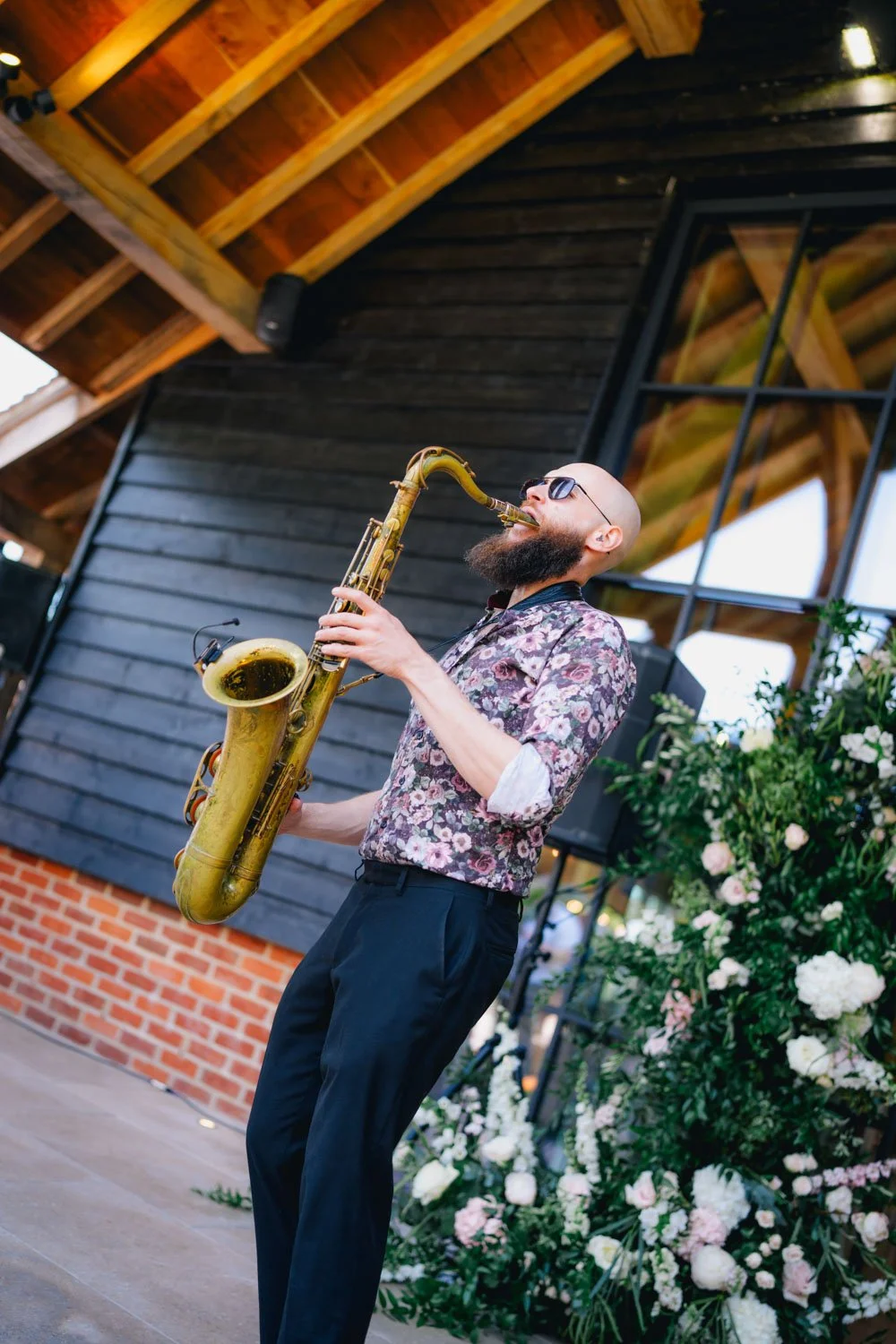 A man with a beard, sunglasses, and wearing a floral shirt playing a saxophone outdoors near a floral arrangement.