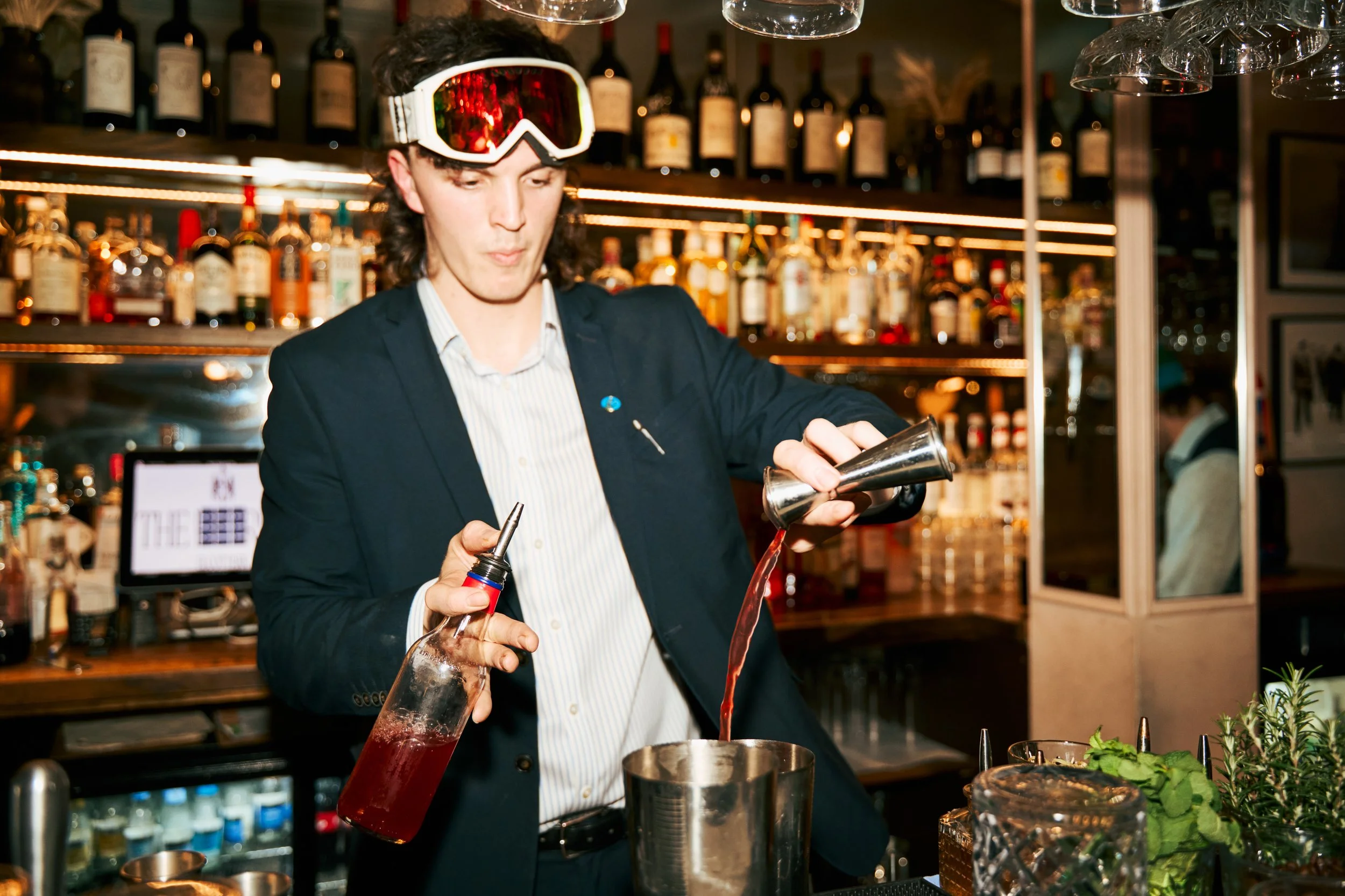 A bartender wearing ski goggles, a dark suit, and a striped shirt pouring a red cocktail from a shaker into a glass at a bar with bottles of alcohol on shelves in the background.
