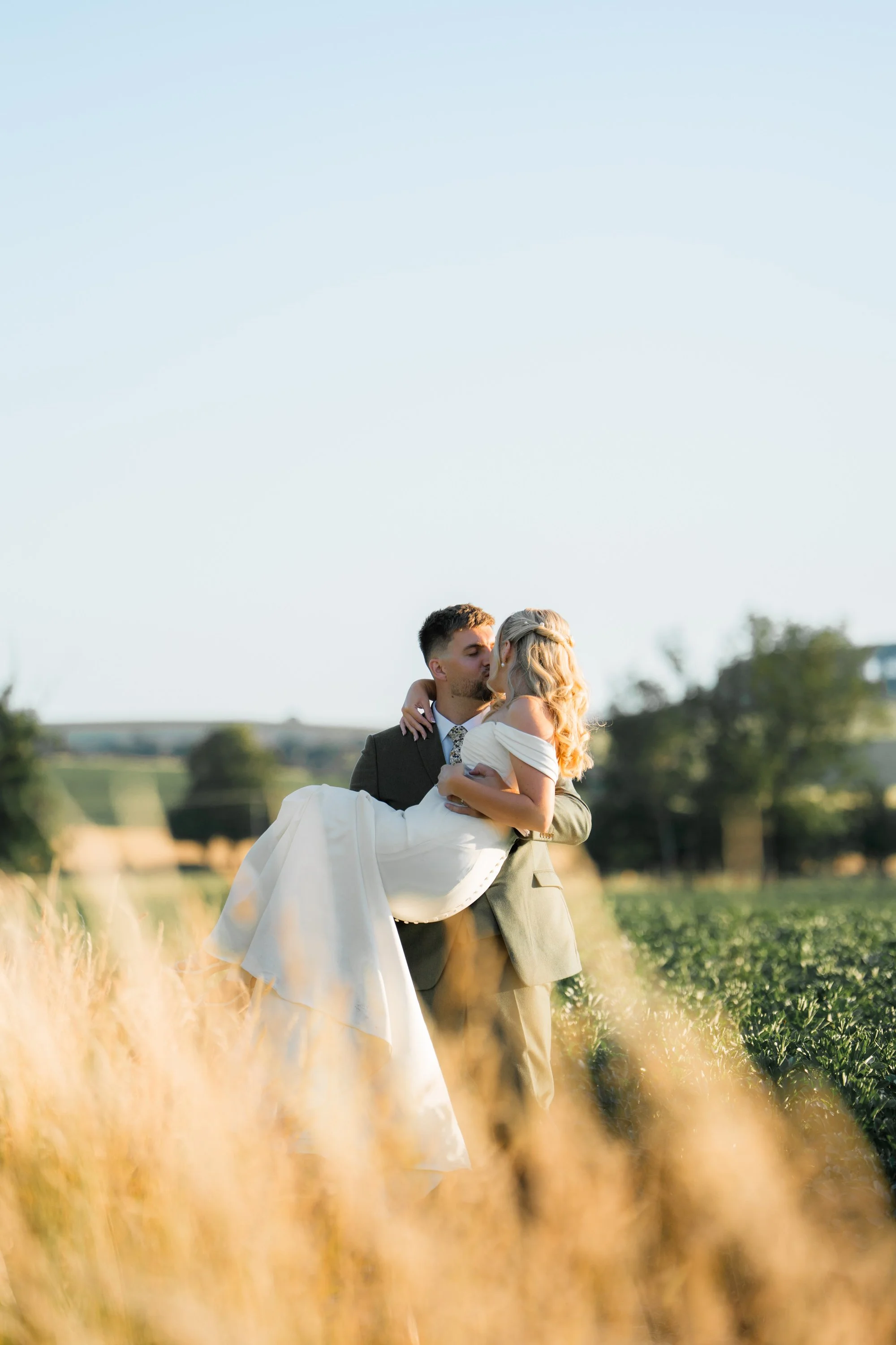 A groom holding a bride in a field with green crops, both dressed formally, during daylight.