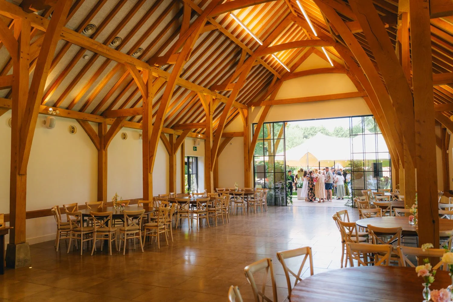 Wooden interior of a spacious event venue with tables and chairs, large open doors leading outside where a crowd of people is gathered under umbrellas.