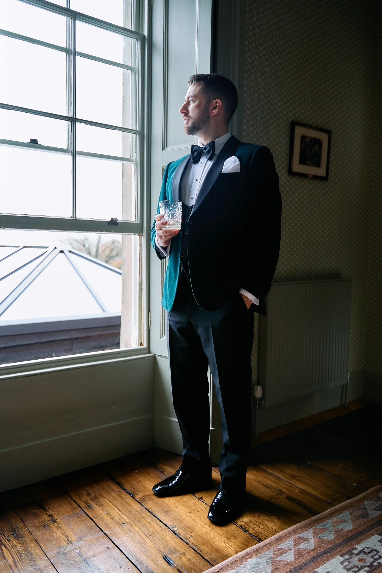 A man dressed in a tuxedo with a bow tie, holding a glass, standing by a window and looking outside.