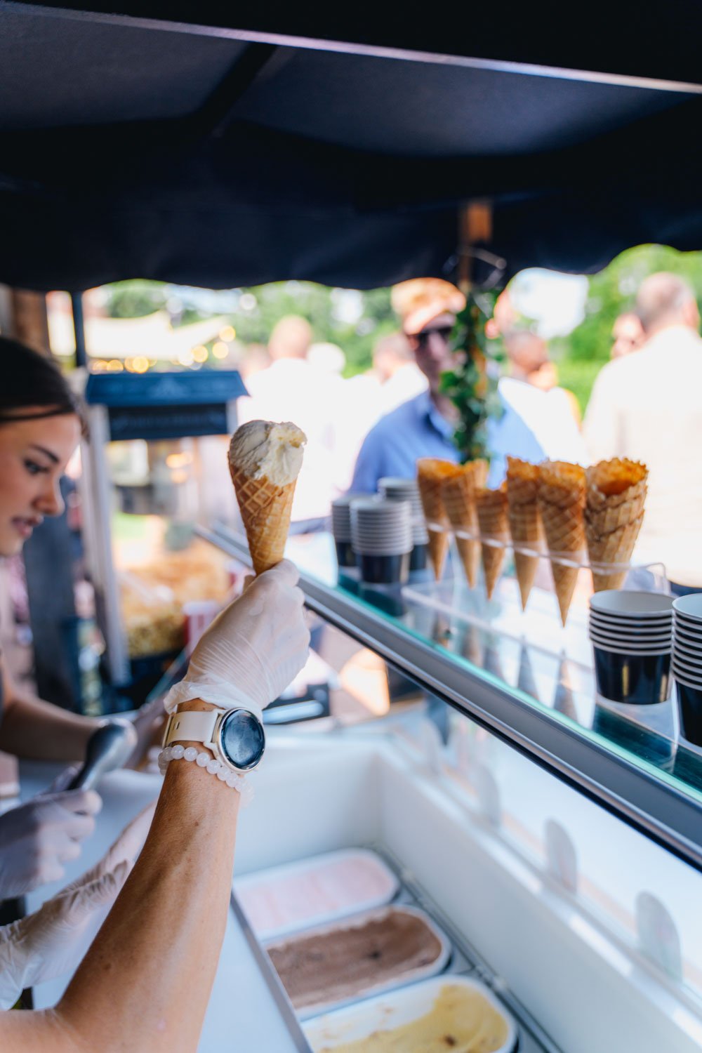 A woman wearing gloves and a watch is holding an ice cream cone with a scoop of vanilla. In the background, a display case contains several more ice cream cones and bowls of ice cream, with a blurry outdoor scene and people.
