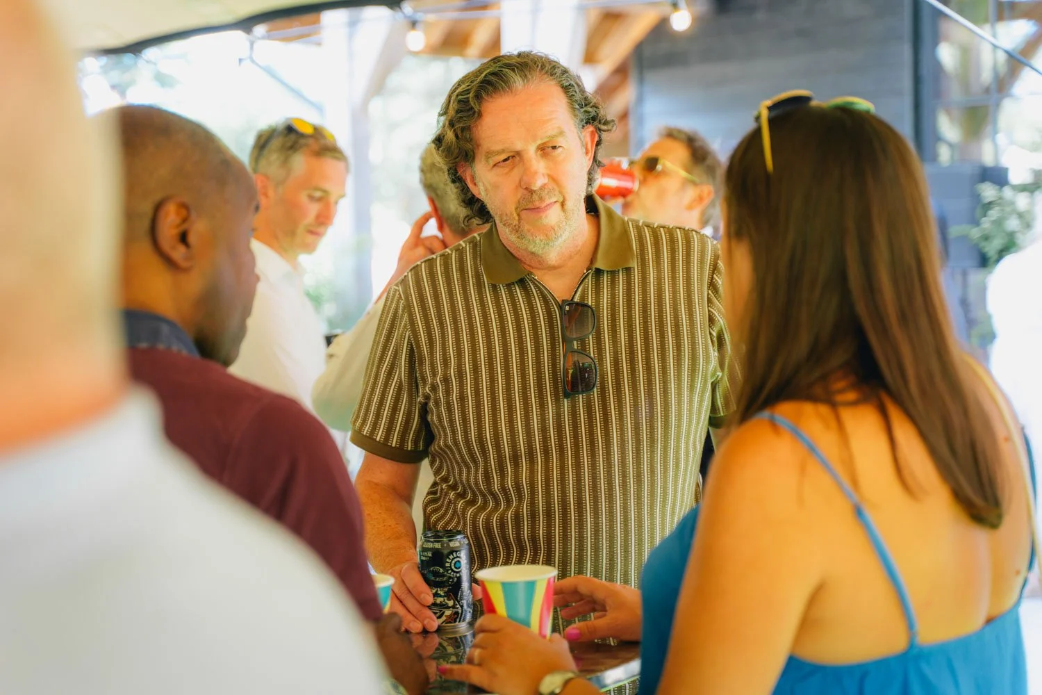 People socializing at a gathering, drinking from cups, in a bright indoor space with wooden accents.
