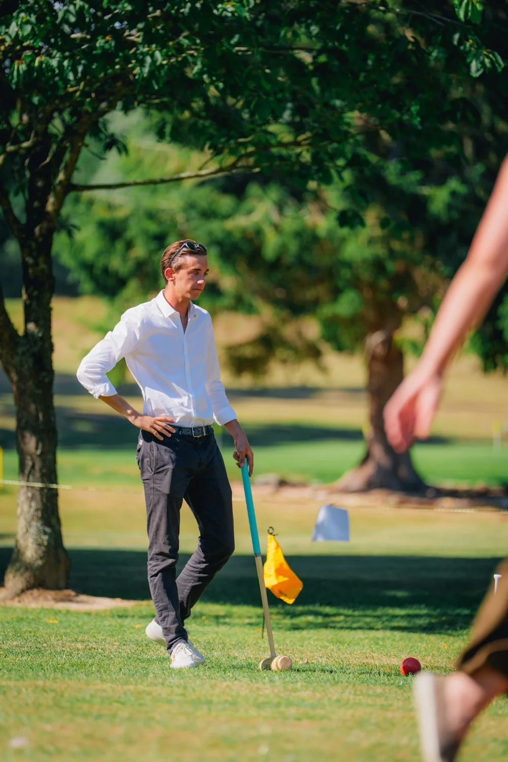 A man in a white shirt and dark pants playing croquet outdoors on a sunny day, with trees and greenery in the background.