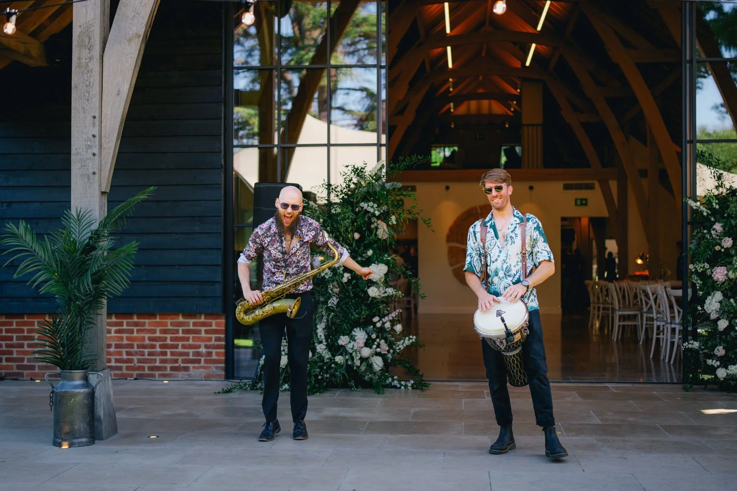 Two musicians performing outdoors in front of a modern barn-style building. One plays the saxophone, the other plays a drum. Both are wearing patterned shirts and sunglasses, smiling and enjoying the event.