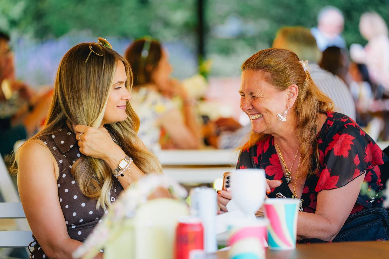 Two women talking and laughing at a gathering, with colorful party cups and decorations on the table in front of them.
