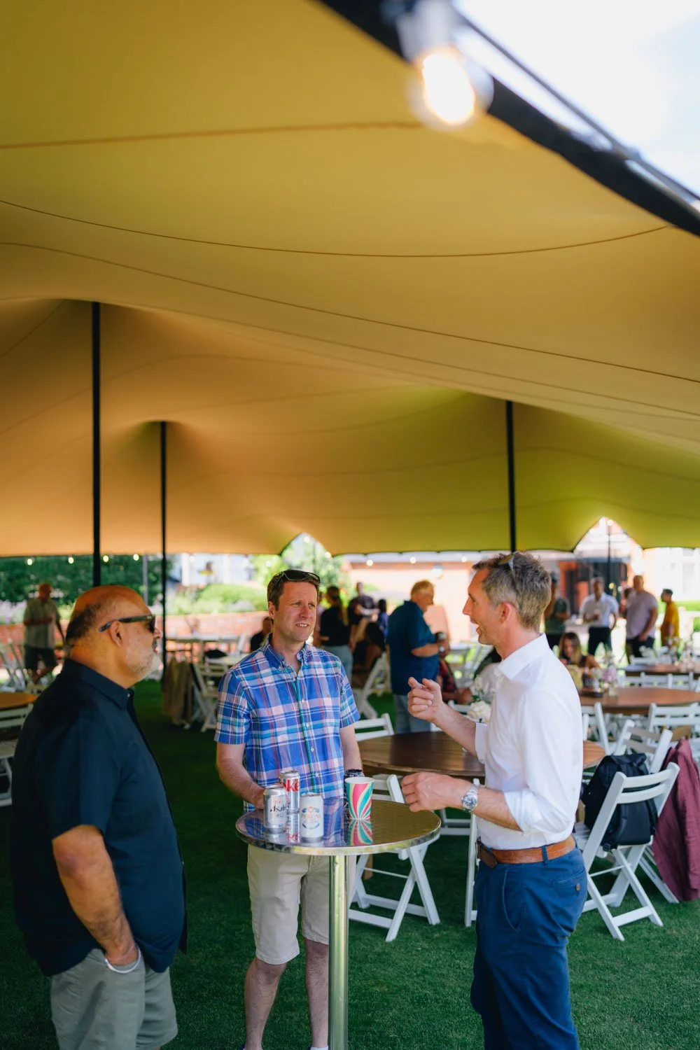 Three men are standing and talking under a large yellow canopy at an outdoor event, with additional people and seating in the background.