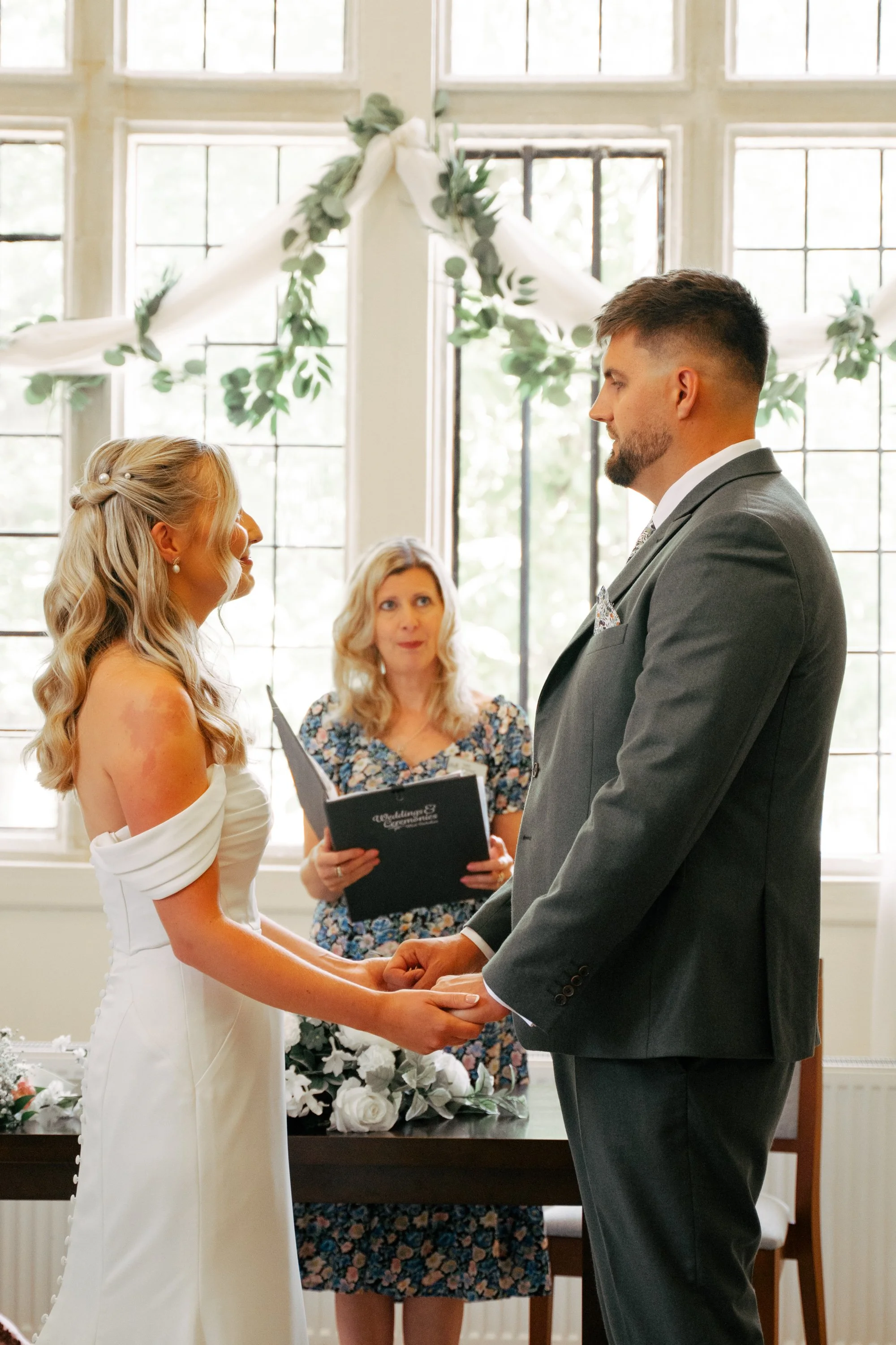 A bride and groom holding hands during their wedding ceremony, standing in front of a large window with natural light, with an officiant and a bridesmaid present.