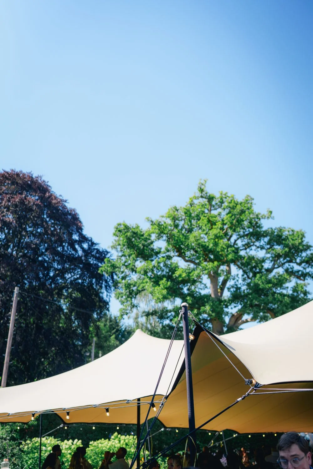 People gathering under a large cream-colored canopy tent outdoors with green trees and a clear blue sky overhead.