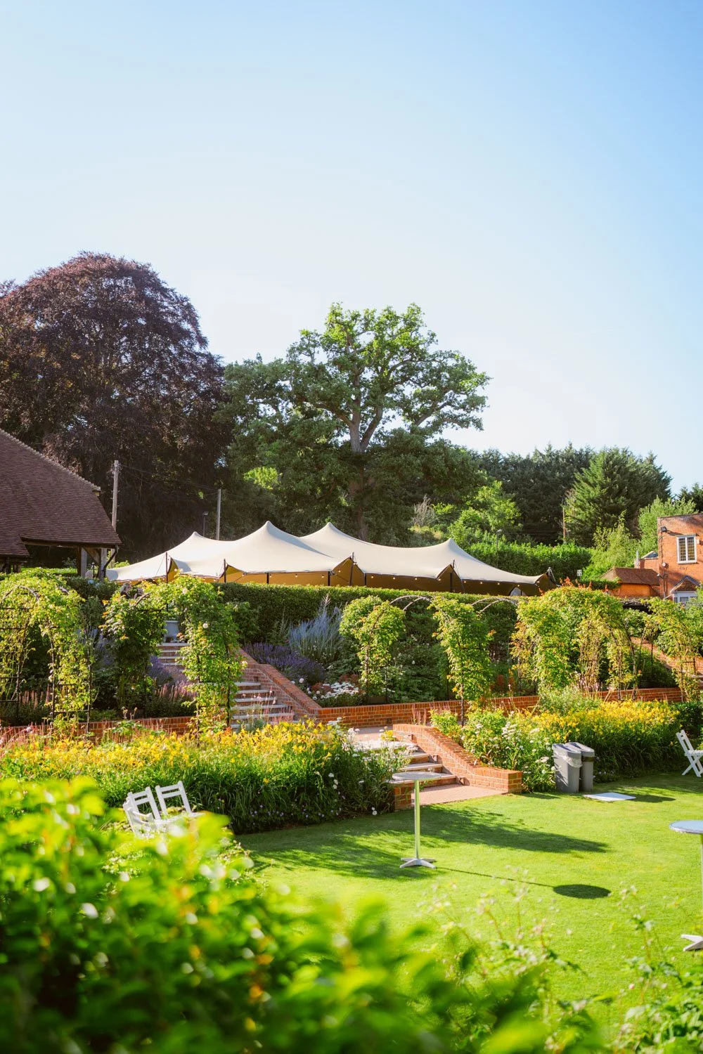 Lush garden with vibrant green foliage, a manicured lawn, a tent with a scalloped edge, brick steps, and trees under a clear blue sky.