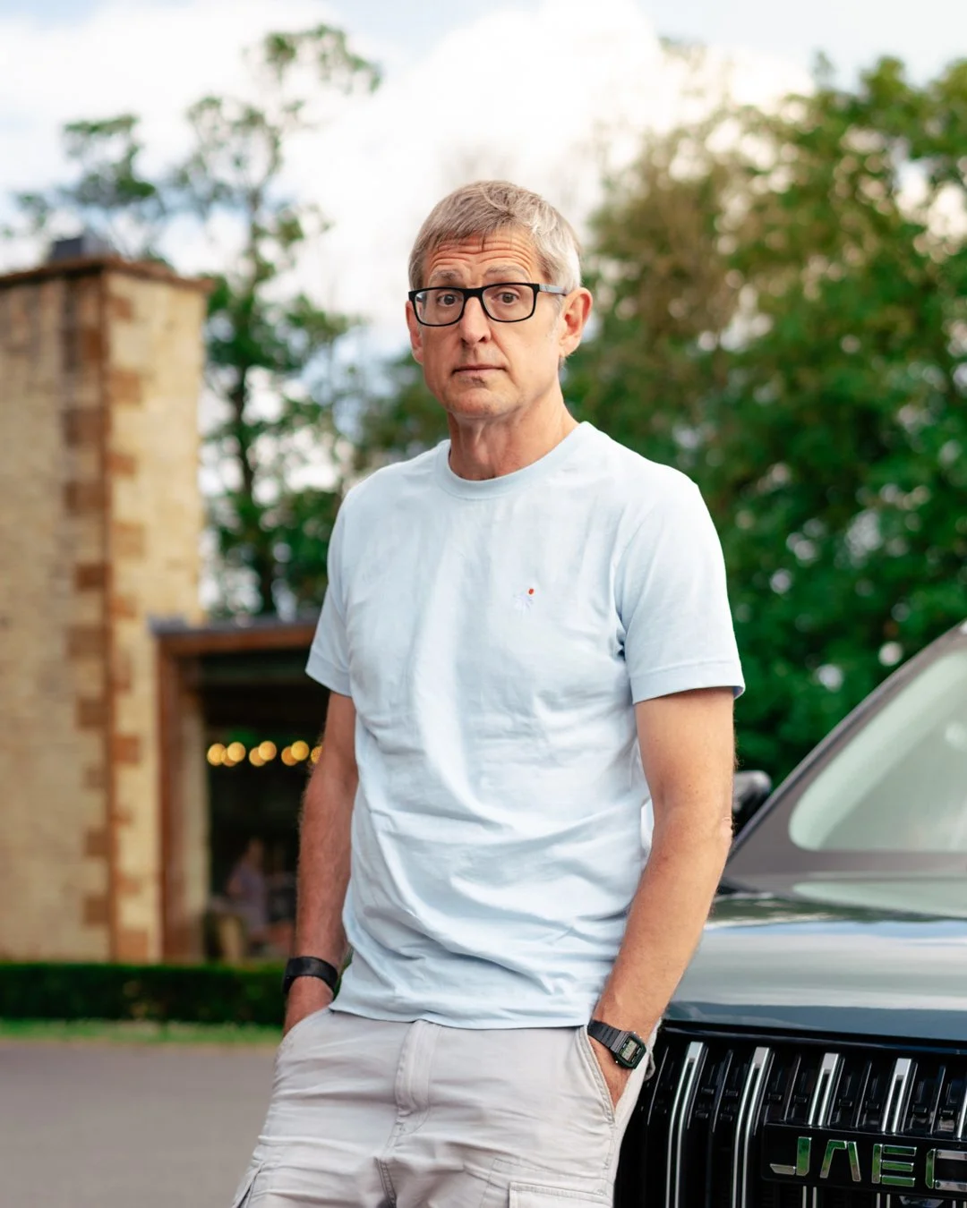 A middle-aged man with gray hair, wearing glasses, a white t-shirt, and beige pants, standing outdoors beside a black SUV, with trees and a brick building in the background.