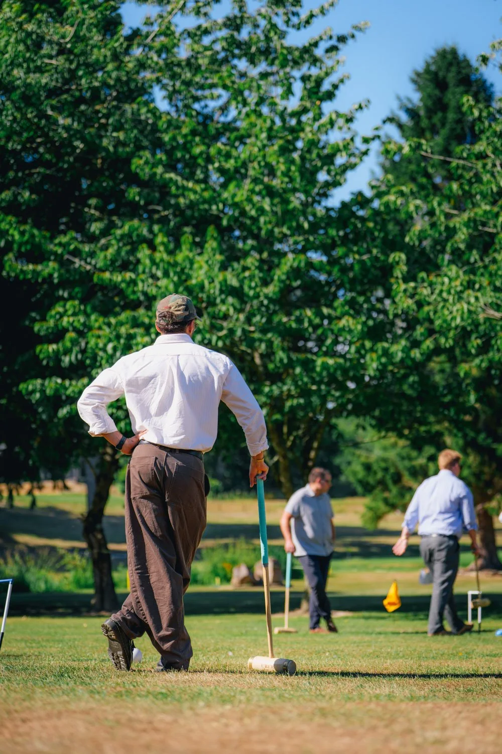A man in a white shirt and brown pants demonstrates playing croquet on a lush green lawn with several other people and trees in the background.