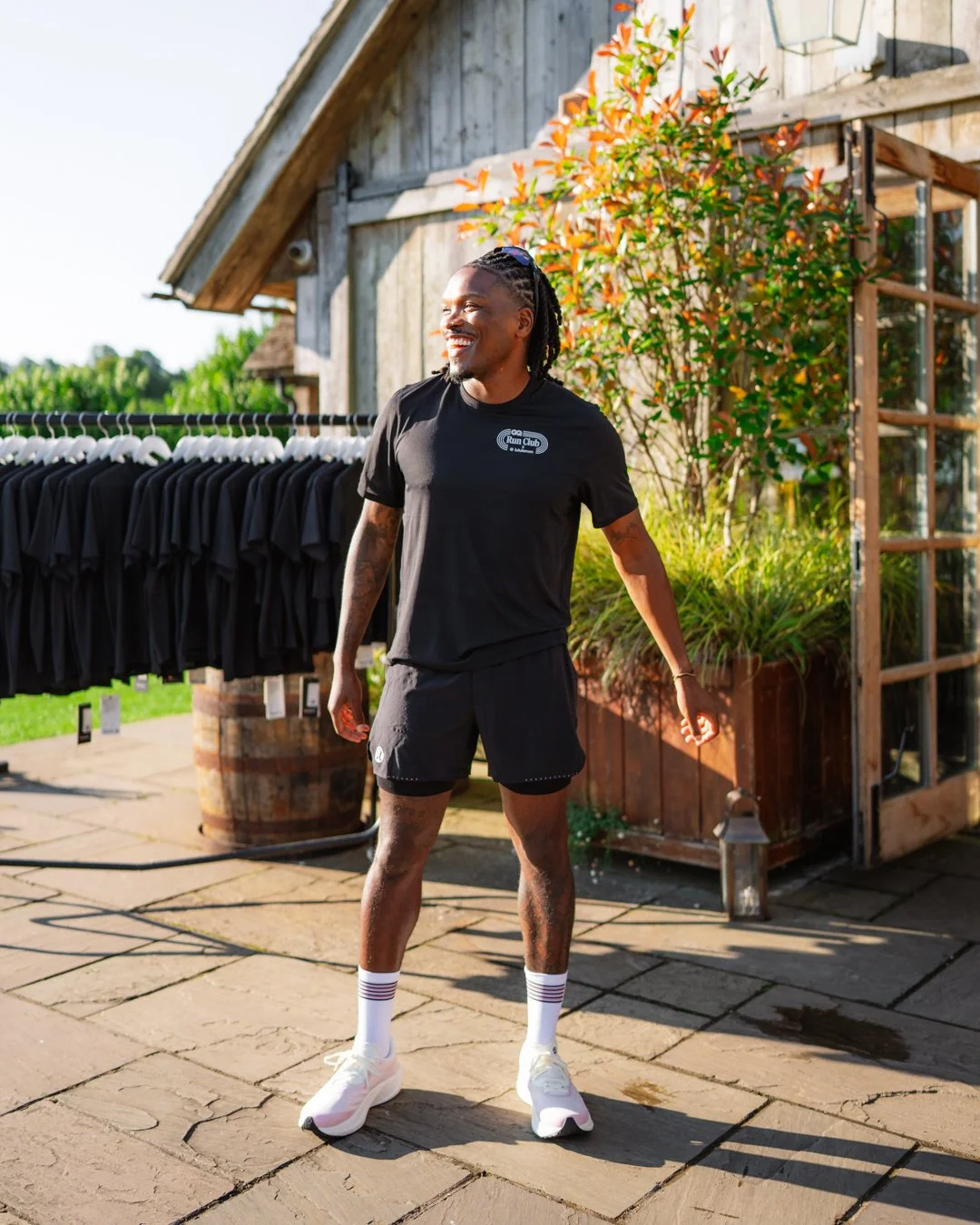 A smiling man in athletic wear standing outdoors on a stone patio, with greenery and a rustic wooden building in the background.