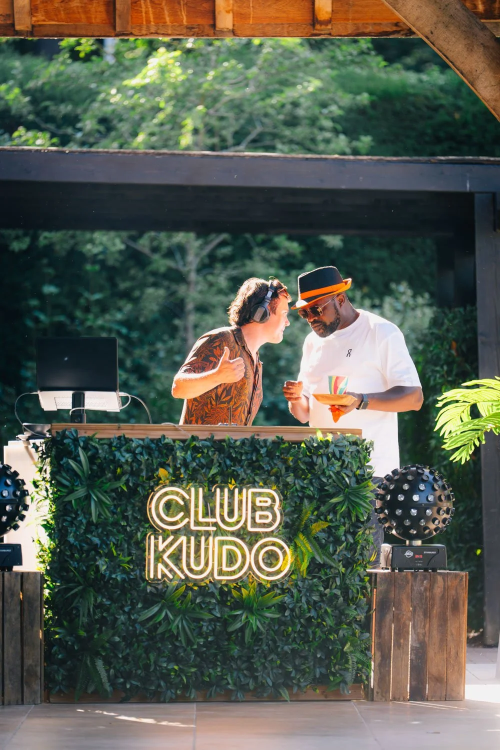 Two men are at a DJ booth with a sign that says "Club Kudo" made of neon light on a leafy green backdrop. One man has headphones and is giving a thumbs up, while the other is holding a plate and talking.
