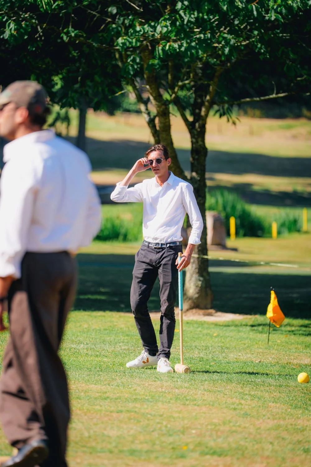 A woman wearing sunglasses and a white shirt standing on a golf course, holding a throw pin and preparing to putt a golf ball while two men, also in white shirts, watch nearby.