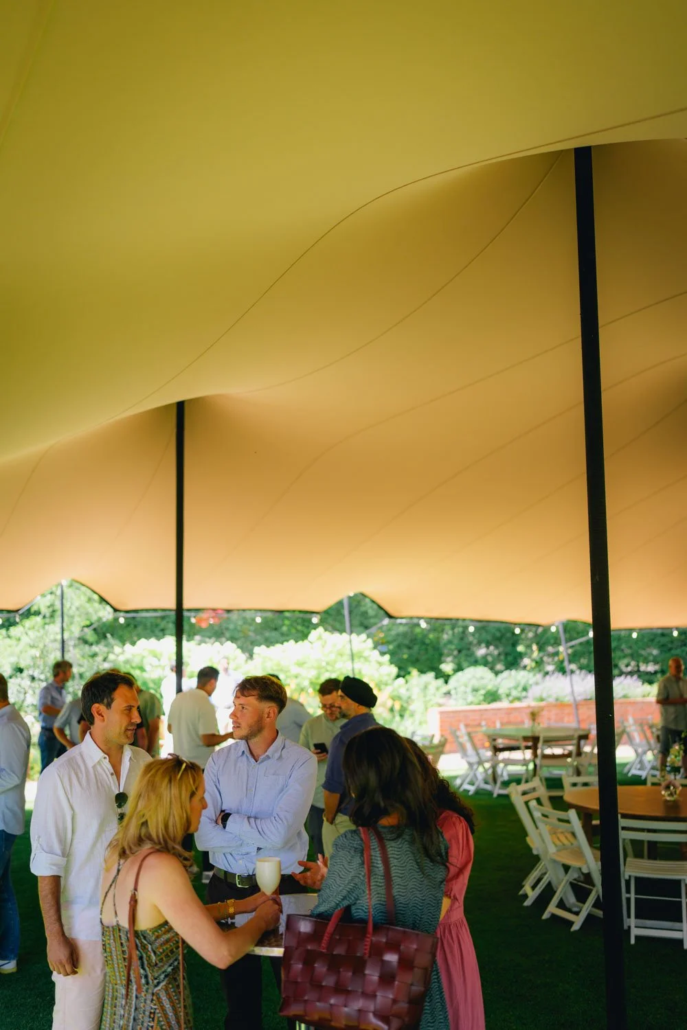 People socializing at an outdoor gathering under a large yellow canopy with green trees and chairs in the background.