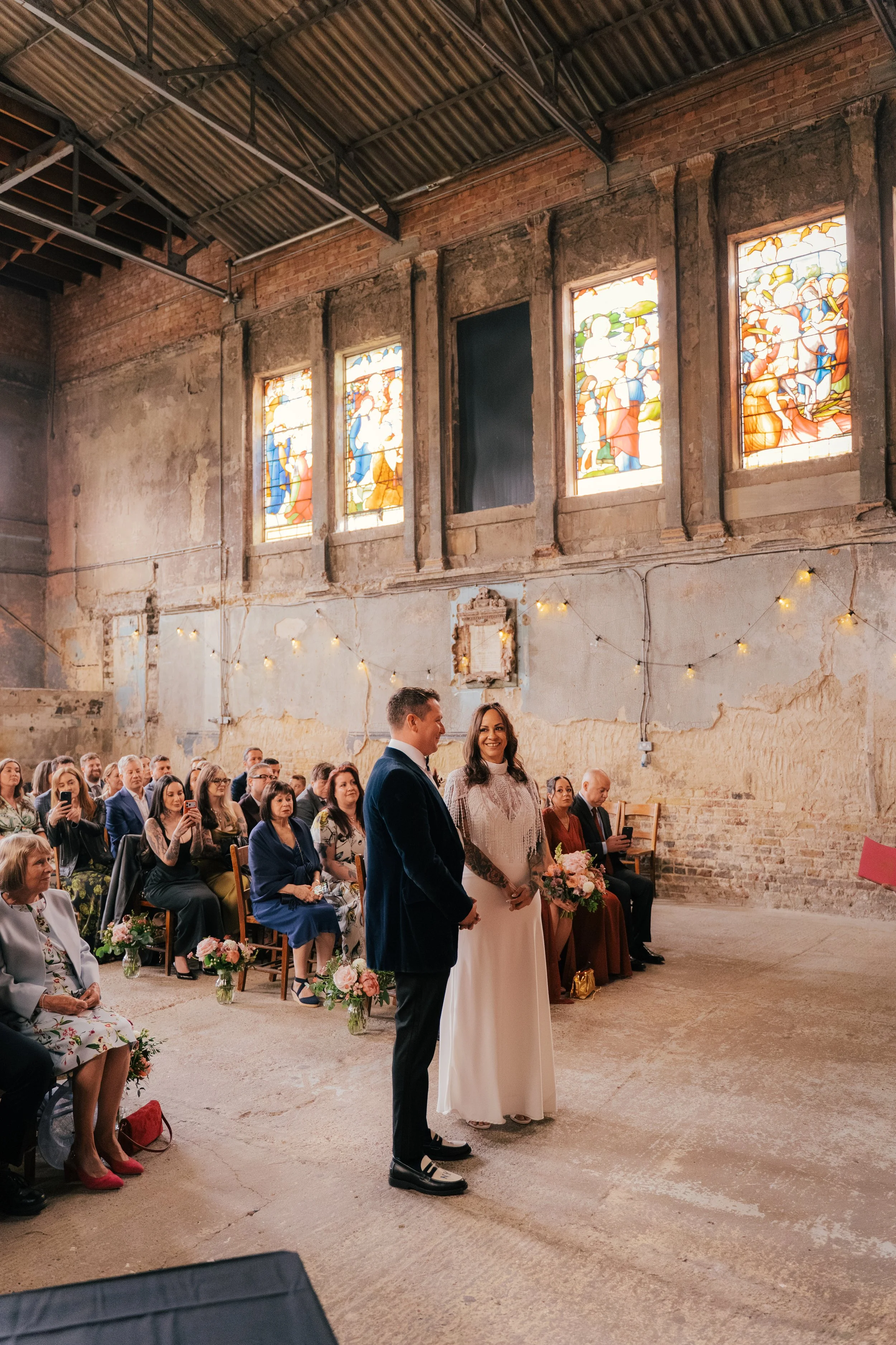 A couple getting married during a wedding ceremony in an industrial-style venue with stained glass windows, exposed brick walls, and string lights. The bride is holding a bouquet of flowers, and both are standing in front of seated guests.