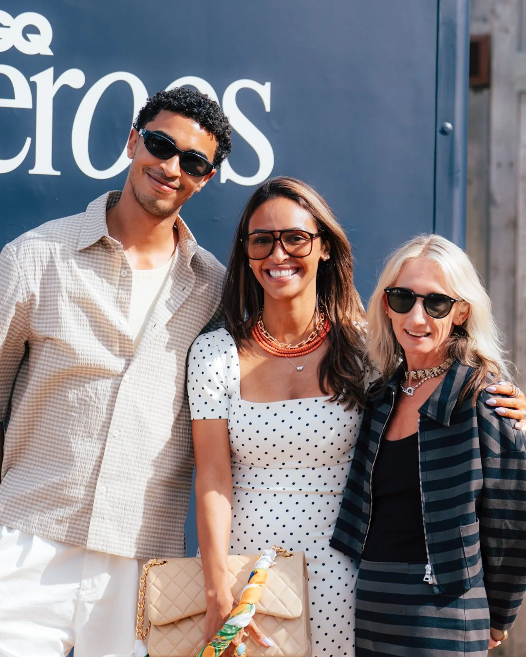 Three friends posing together outdoors in front of a sign that says 'cross' in large white letters, smiling and wearing sunglasses.
