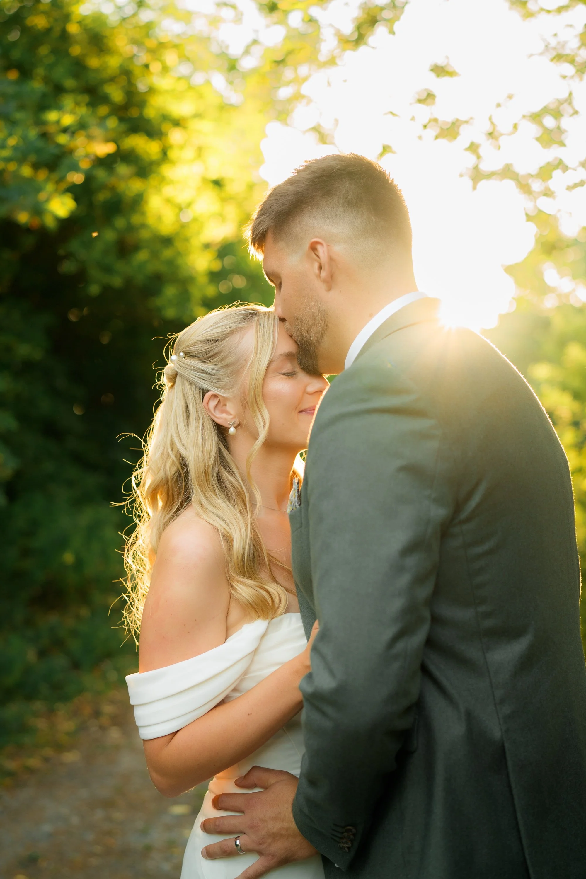 A couple on their wedding day outdoors, standing close with the groom kissing the bride's forehead, illuminated by the sun in the background.