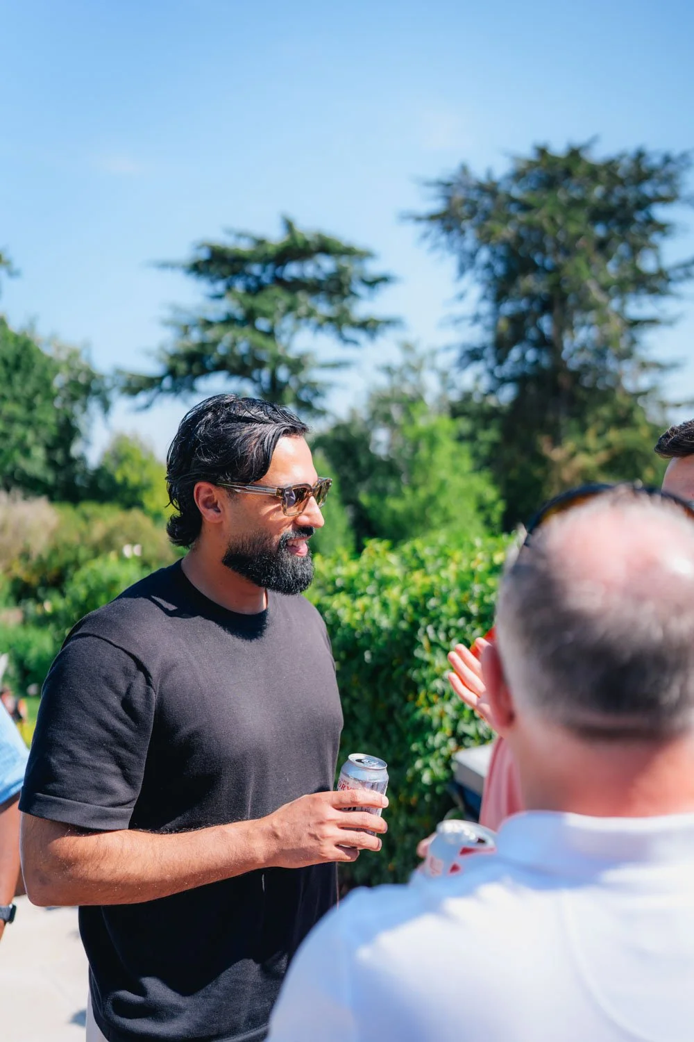 A man with dark hair, sunglasses, and a beard holding a soda can, engaging in conversation outdoors with a group of people on a sunny day, green trees in the background.