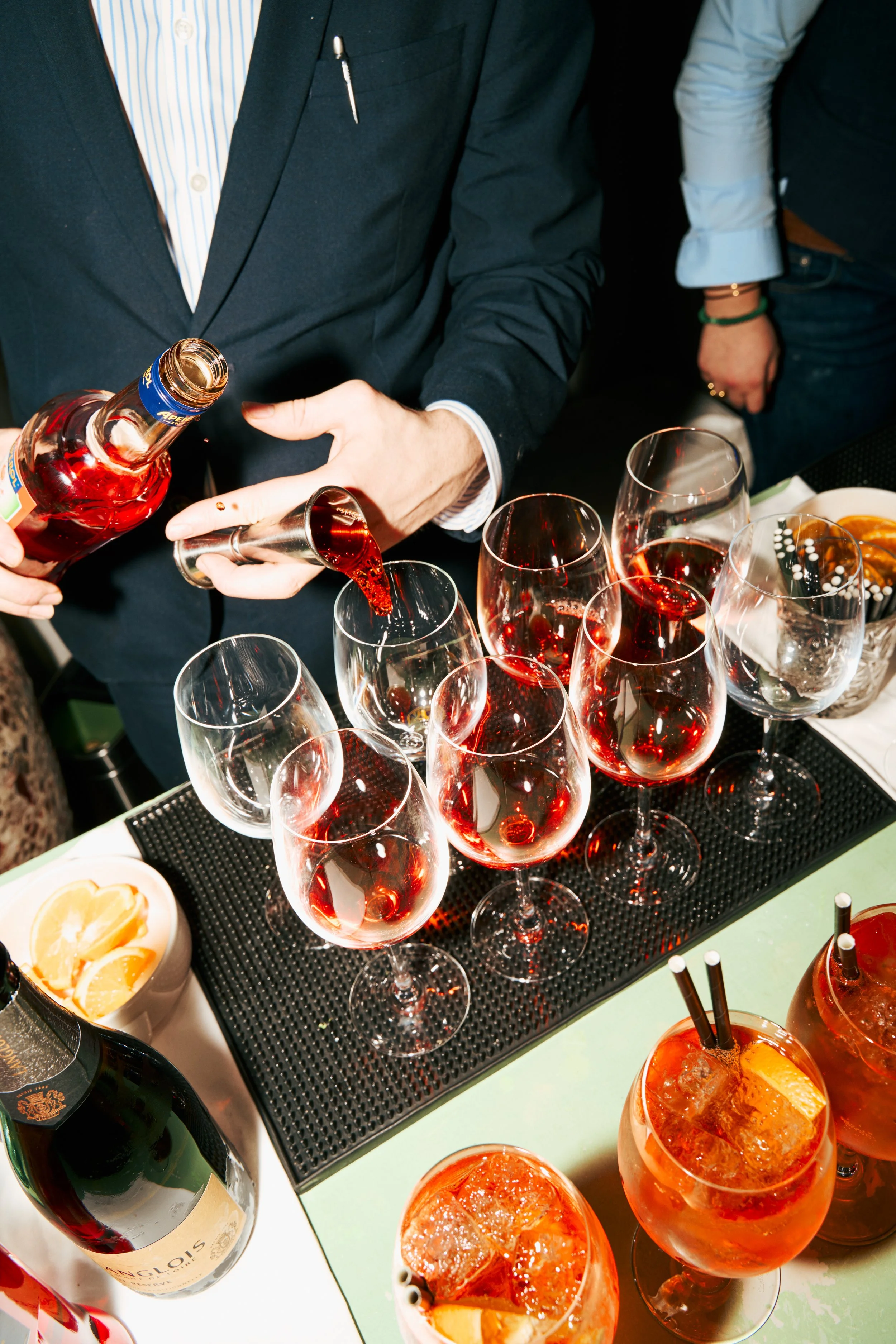 A person in formal attire pouring red wine into glasses at a gathering, with additional glasses and drinks on the table.