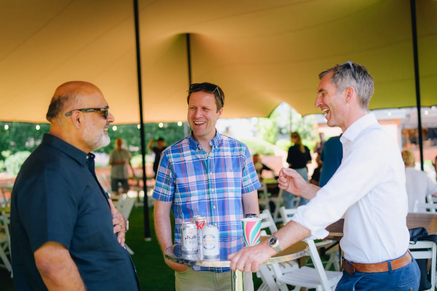Three men having a conversation and smiling during an outdoor gathering under a canopy, with drinks on a small round table in front of them.