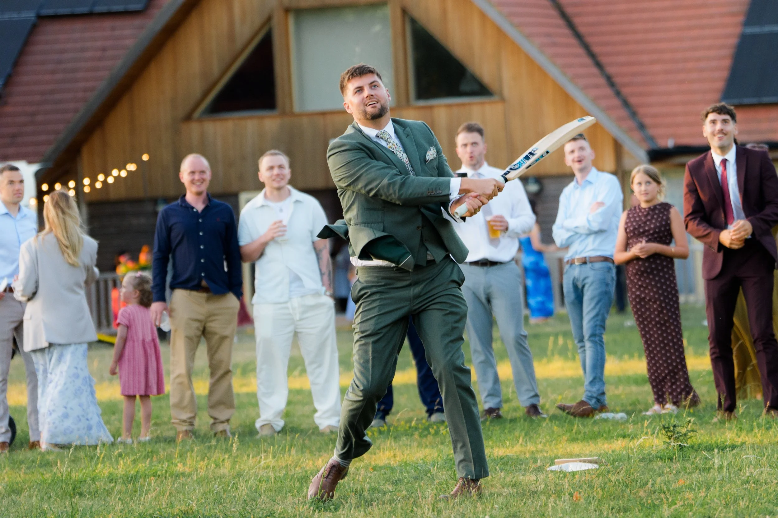 A man in a green suit and floral tie is playing cricket at an outdoor wedding reception, with guests watching and smiling in the background.