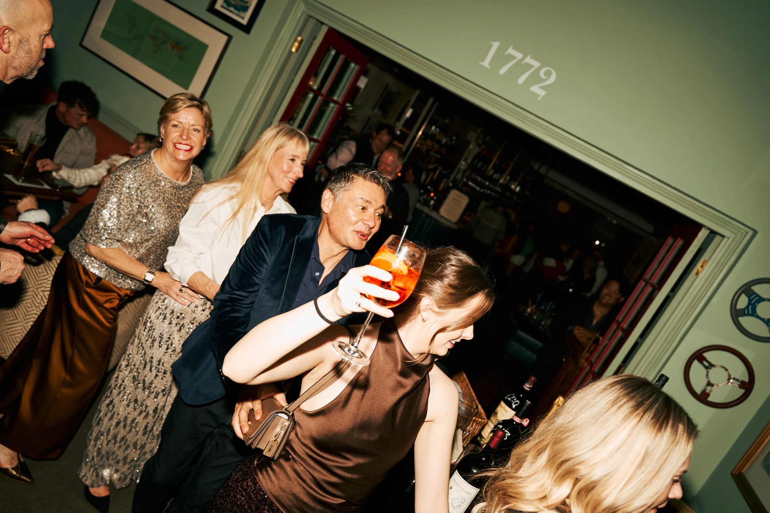 People standing in line at a bar or restaurant, with a woman in the foreground holding a drink and women in the background smiling and chatting, with the year 1772 painted on the wall above.