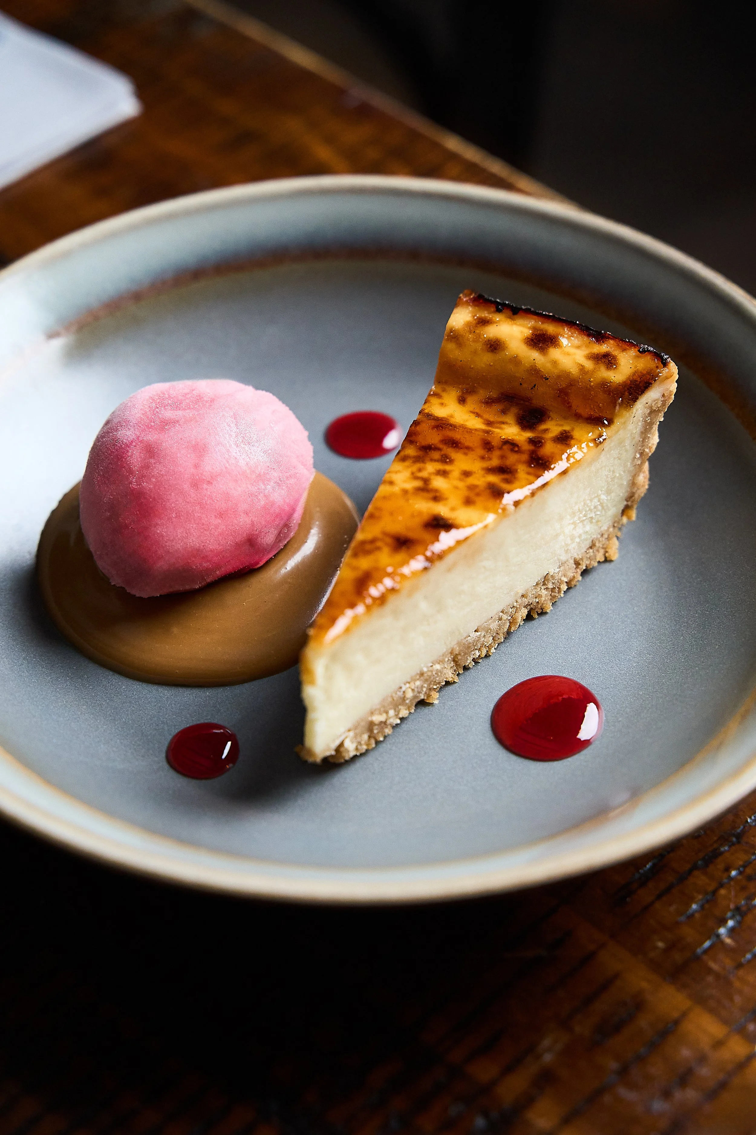A dessert plate featuring a slice of cheesecake with a caramelized top, a pink sorbet on a chocolate sauce bed, and small red fruit sauce dots.