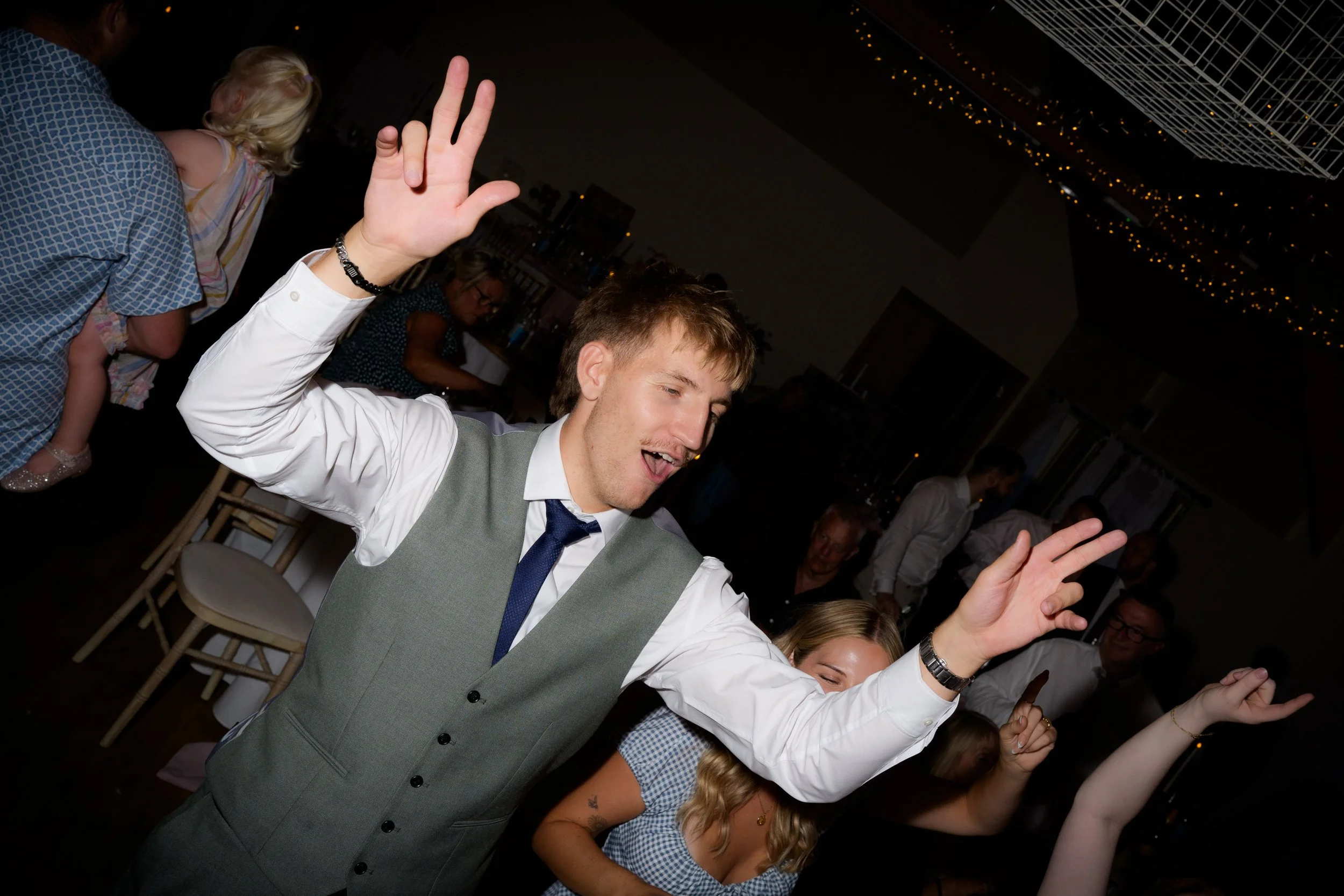 Young man in a gray vest and white shirt dancing with his eyes closed at a party, with other guests dancing in the background.