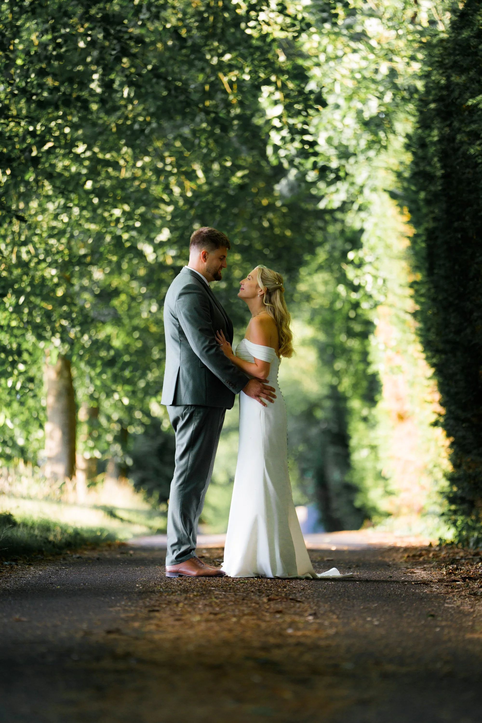 A bride and groom standing close together on a tree-lined pathway, gazing at each other during a wedding photoshoot.