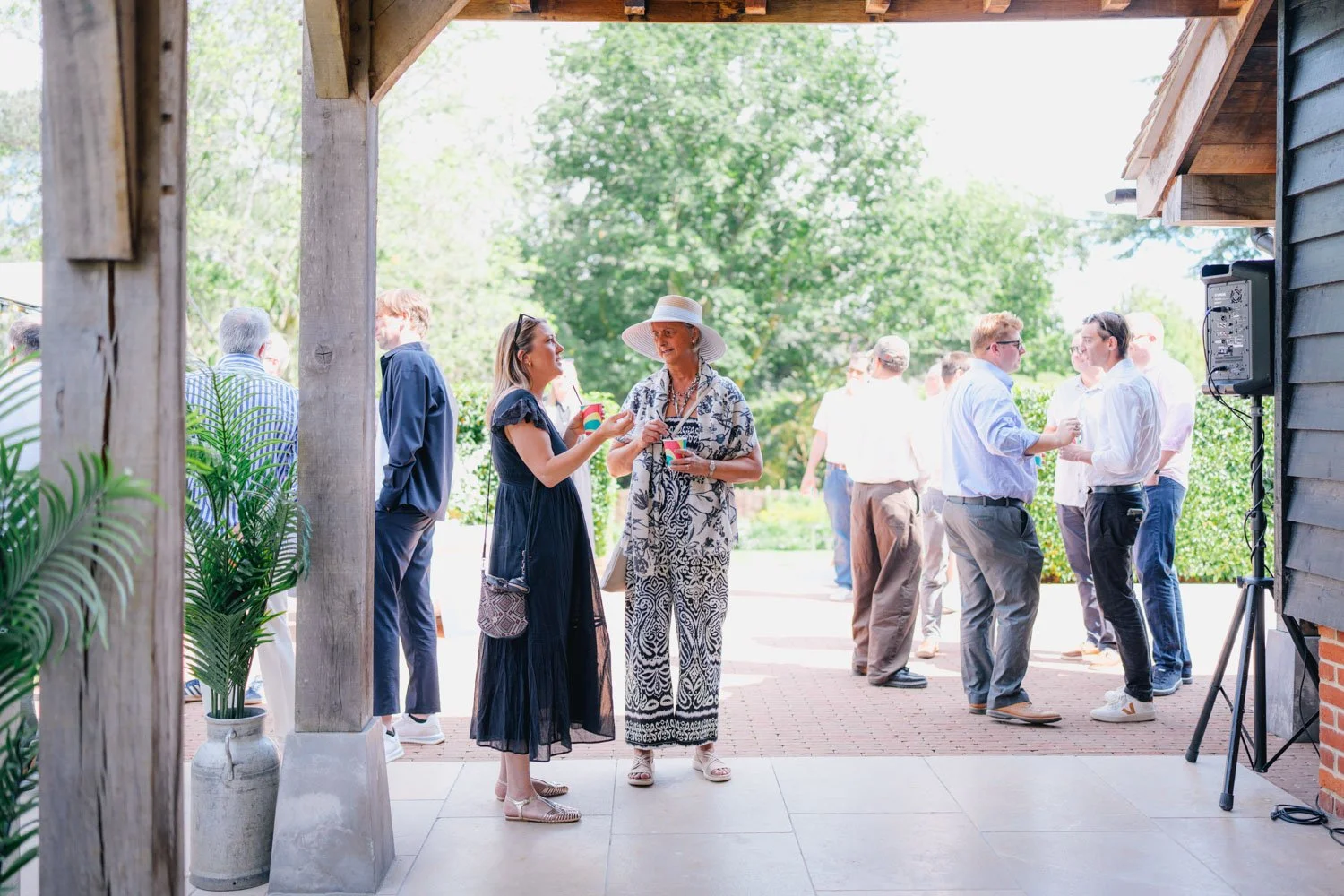 People socializing outdoors on a sunny day, with a woman in a black dress and a woman in a patterned dress wearing a large sun hat holding drinks, and others in the background engaged in conversation.
