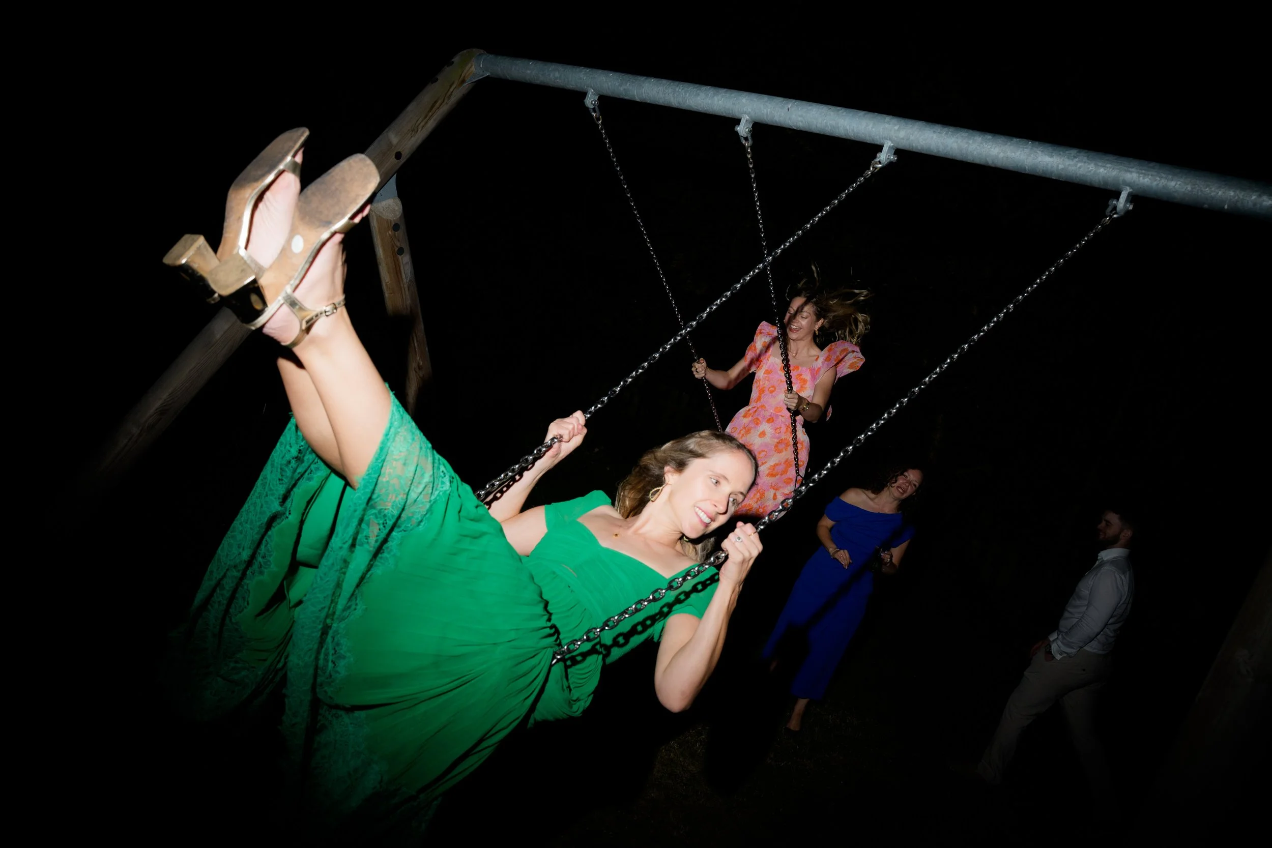 Group of women in colorful dresses swinging on a playground swing set at night.