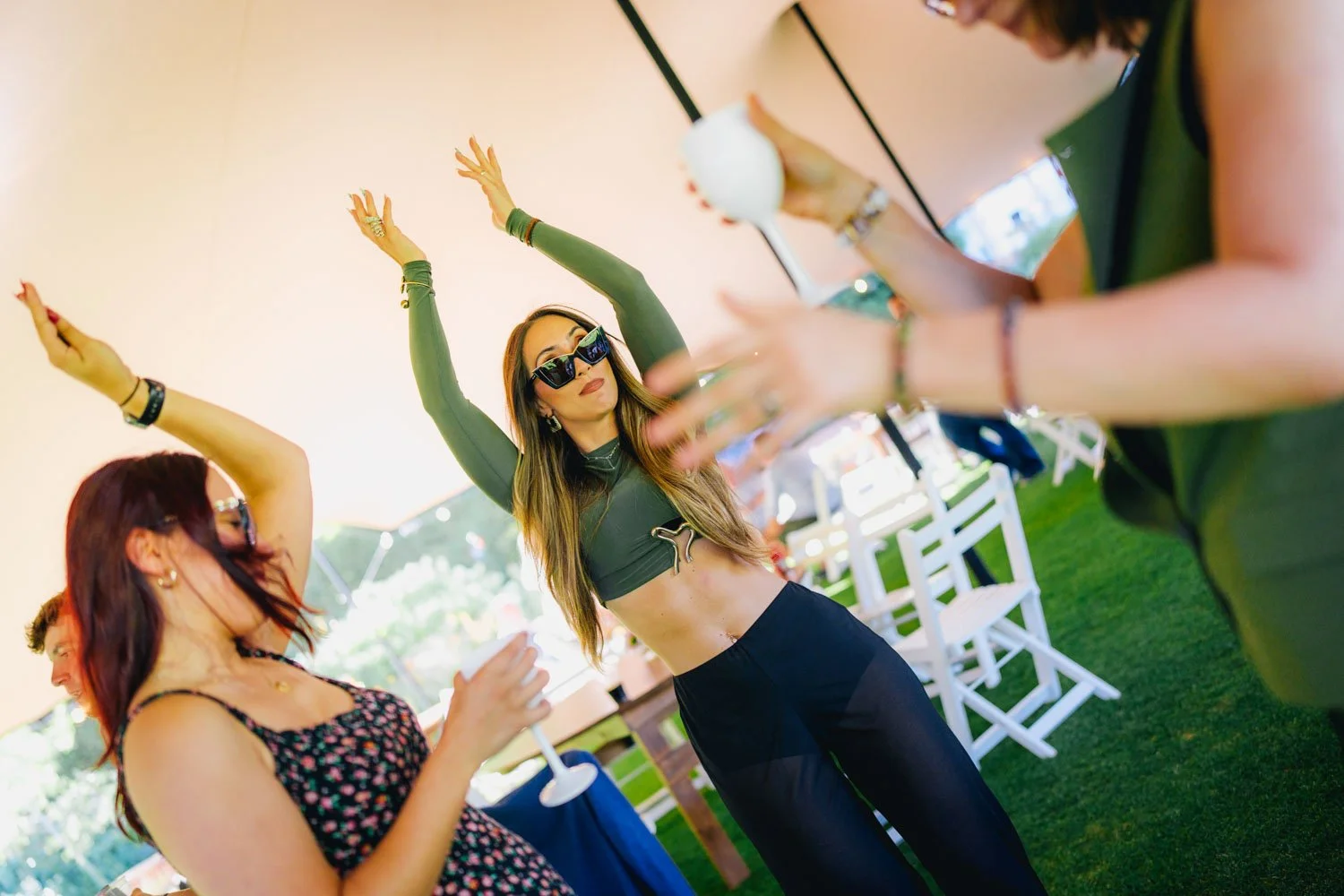 Young woman with long hair wearing sunglasses, a green crop top, and black pants dancing with her arms raised under a large outdoor tent at a party. Other women are nearby, some holding drinks, and white chairs are set up on grass.