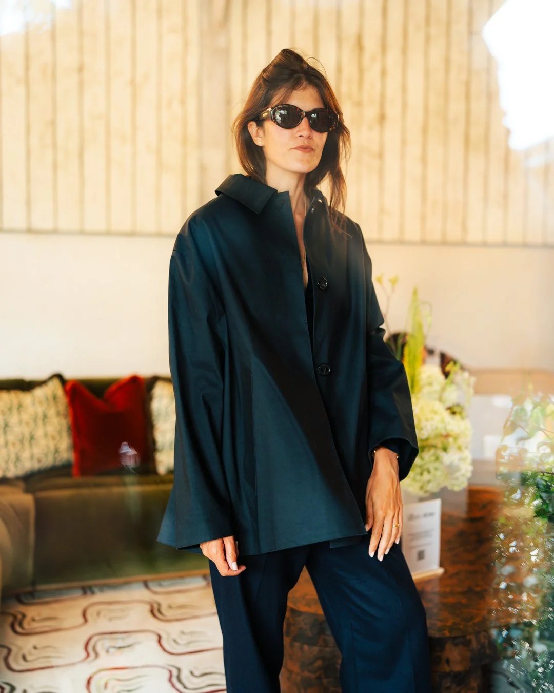 A woman wearing a black coat and sunglasses standing indoors with a blurred background of a wooden wall, floral arrangement, and a vintage-style sofa.
