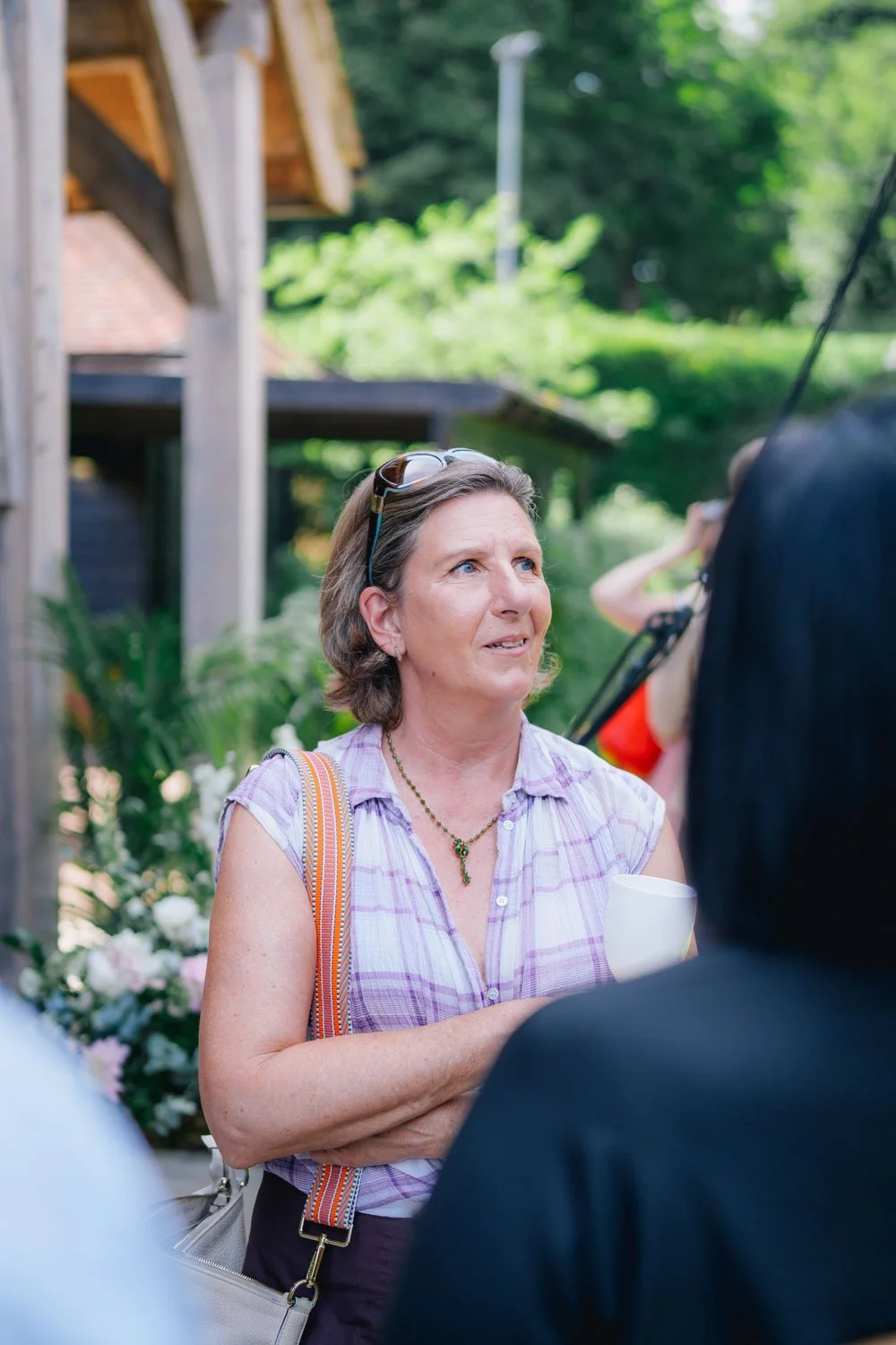 A woman with short gray hair, wearing a purple plaid shirt, glasses on her head, and a necklace, is holding a white cup and looks slightly puzzled or concerned while conversing with someone outside of the frame. There are greenery, a wooden structure