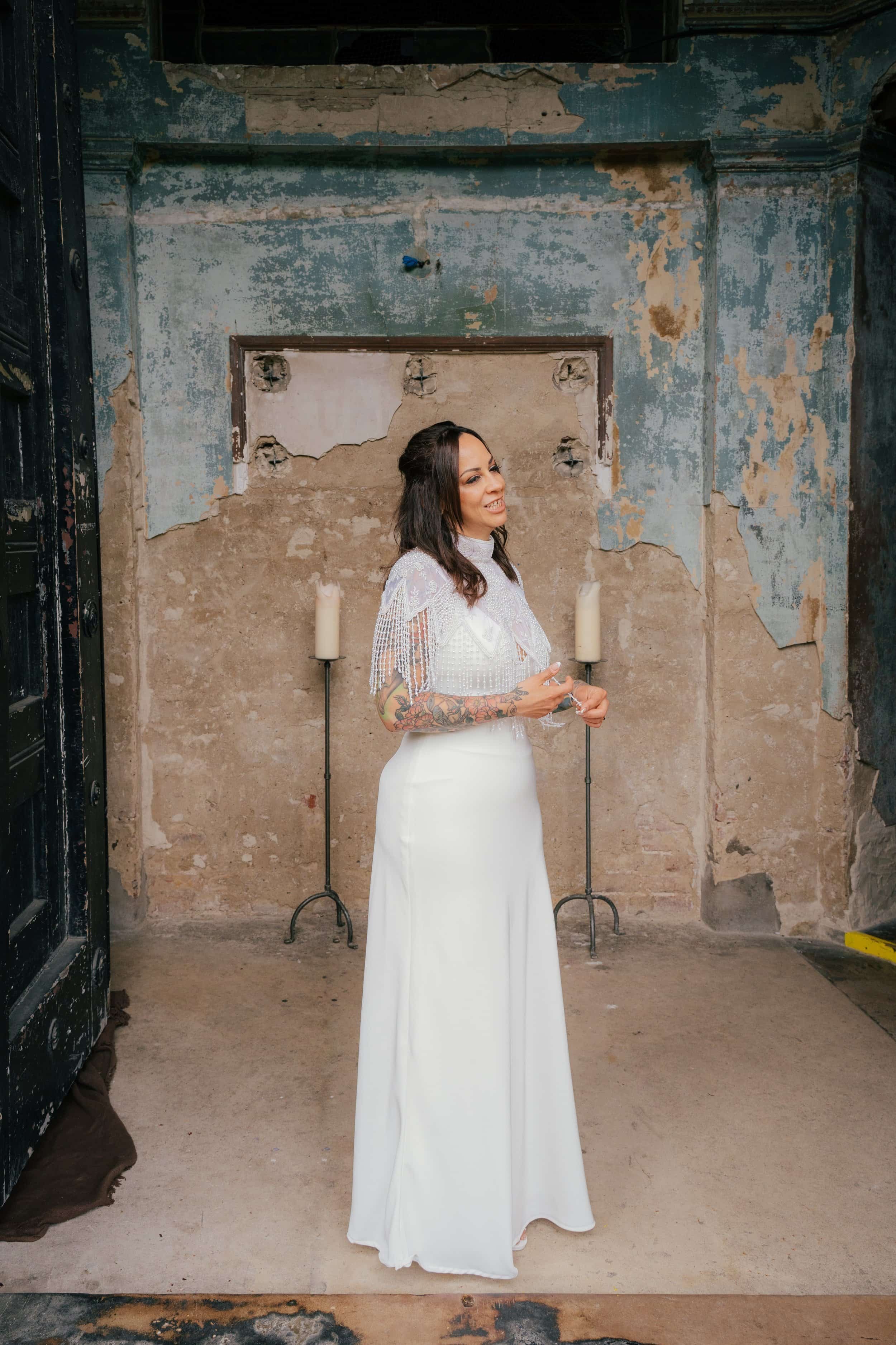 A woman in a white dress standing in front of a weathered, peeling wall with two candles on candleholders behind her, taken by UK wedding photographer, Alex Gell